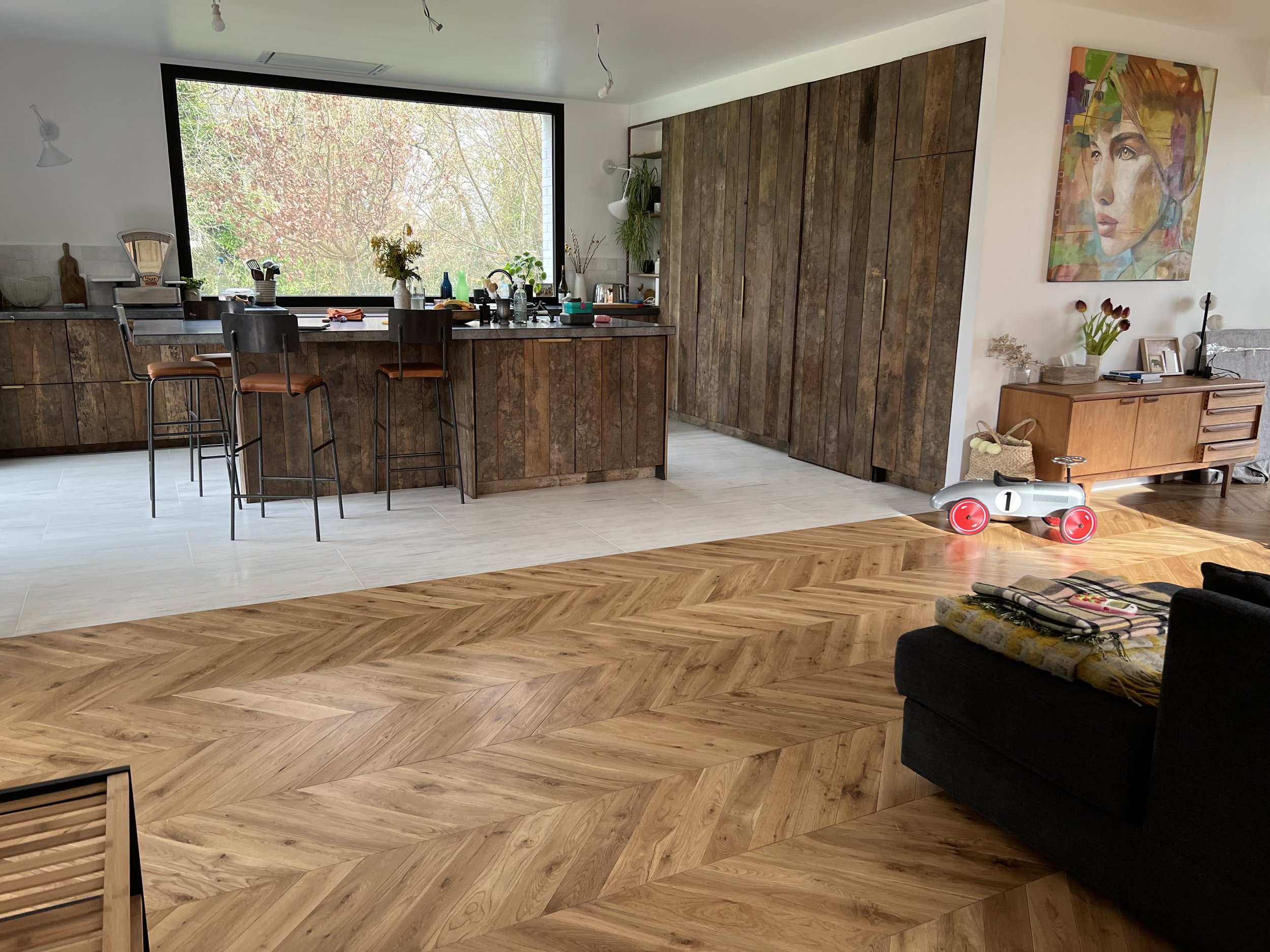 Cuisine ouverte moderne avec îlot central en bois, tabourets, grandes fenêtres, parquet à chevrons et coin salon décoré.