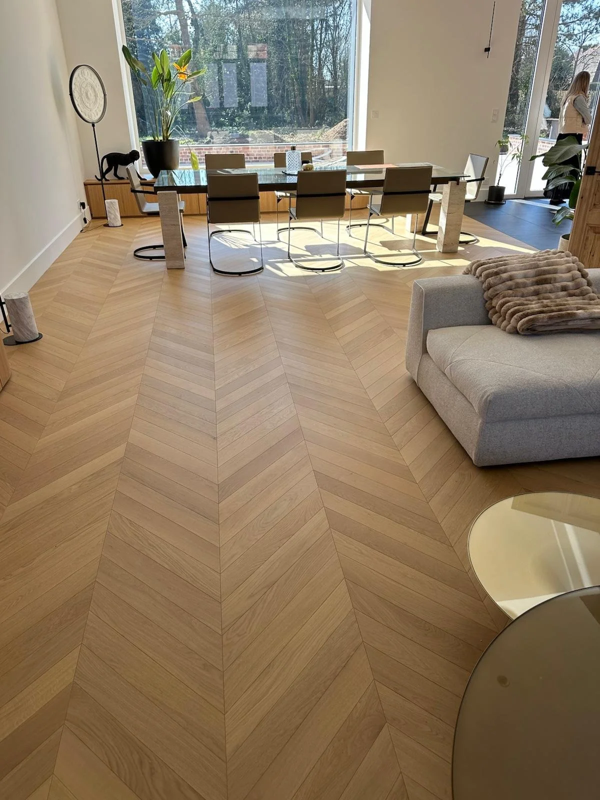 Salle à manger moderne avec table en verre, chaises contemporaines, parquet à chevrons, grandes baies vitrées et canapé beige.