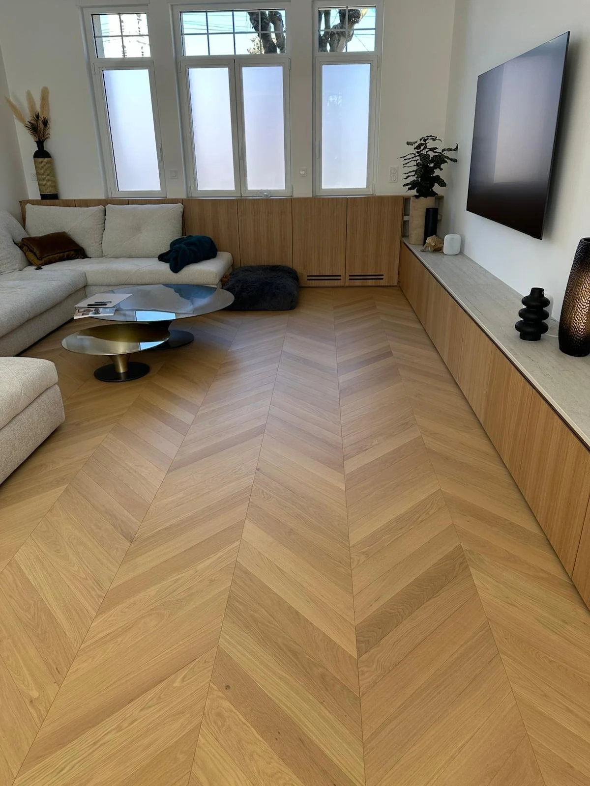 Salon moderne avec canapé d’angle, parquet en chevrons, meuble TV en bois, téléviseur mural et grandes fenêtres.