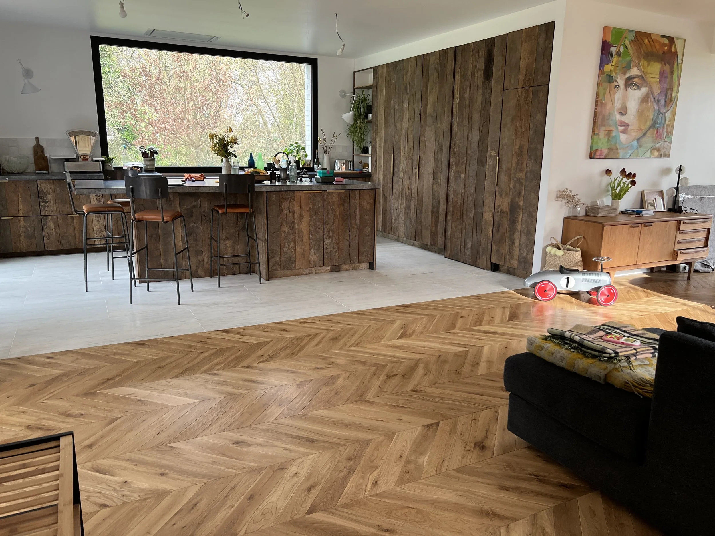 Cuisine ouverte moderne avec meubles en bois brut, îlot central, parquet à chevrons, grande fenêtre et espace salon