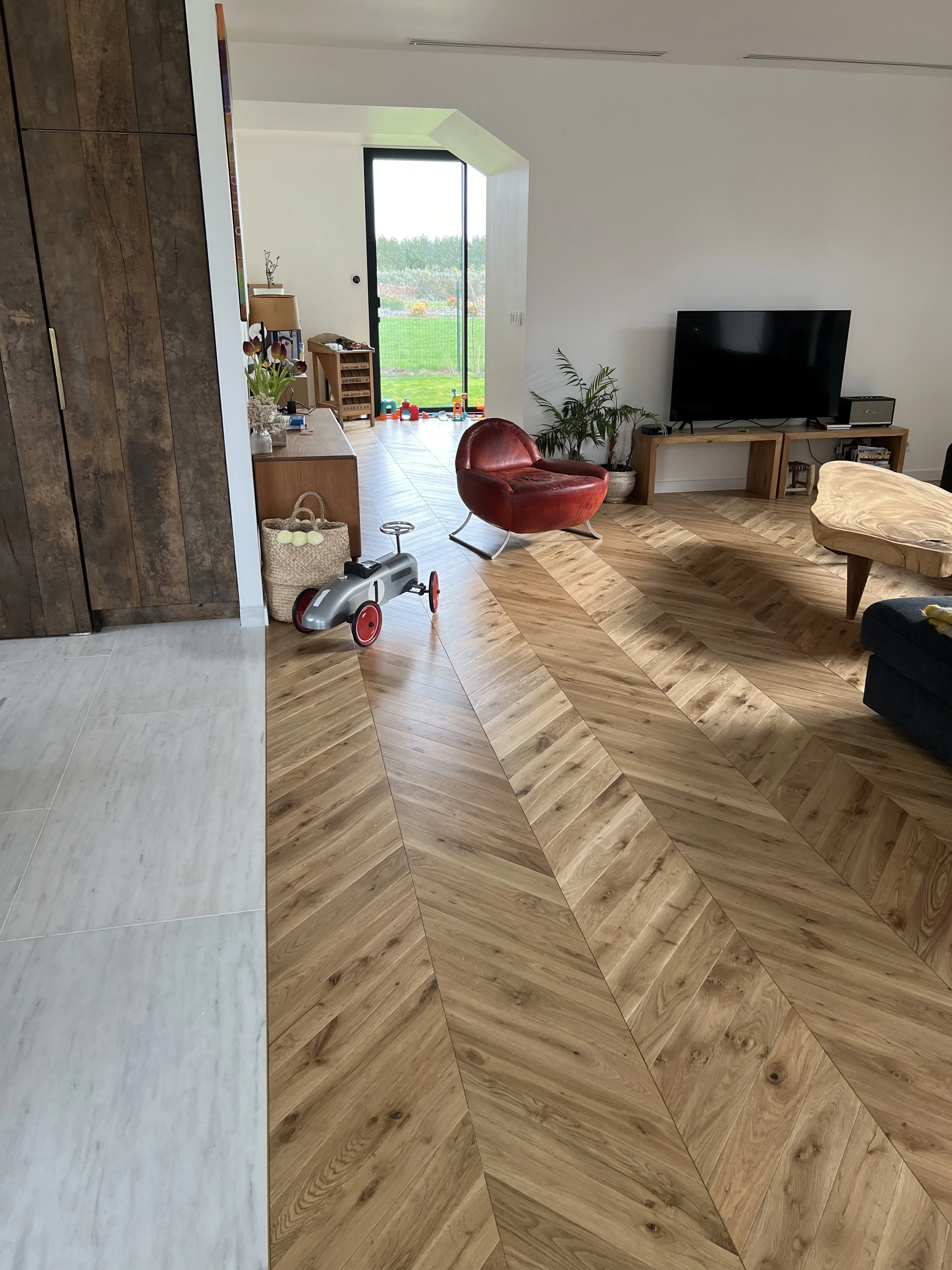 Salon moderne avec téléviseur, fauteuil rouge, table basse en bois, plantes et grande baie vitrée donnant sur un jardin.