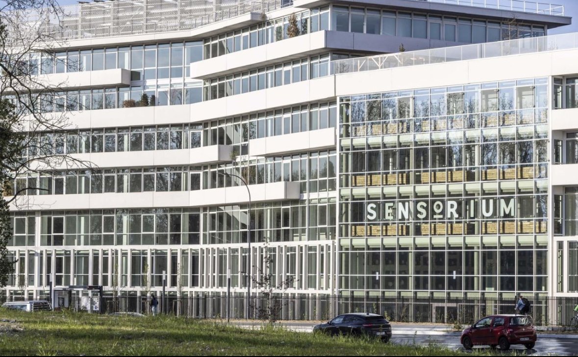 Façade moderne du Sensorium, bâtiment vitré à plusieurs étages avec voitures au premier plan.