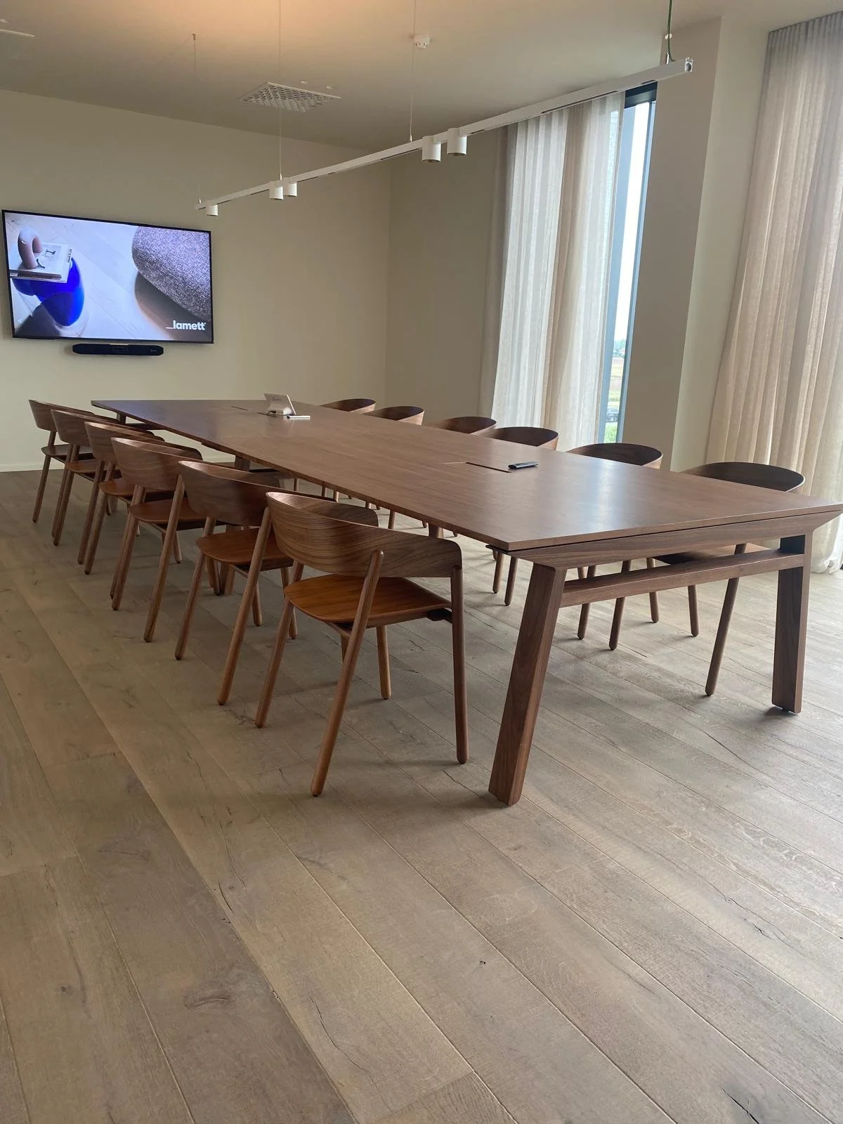 Salle de réunion moderne avec grande table en bois, chaises, écran mural et fenêtres hautes avec rideaux.
