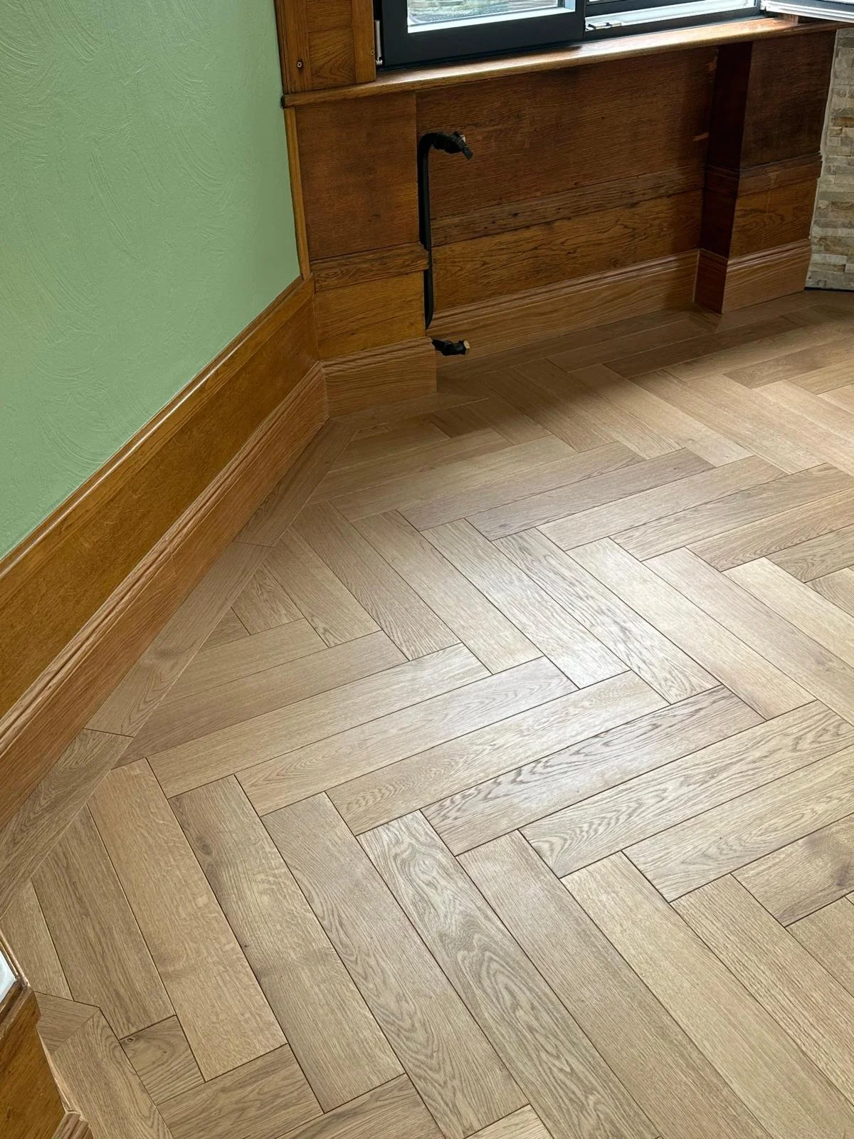 Parquet en bois clair posé en chevrons dans une pièce aux boiseries murales et aux raccords de plomberie apparents.