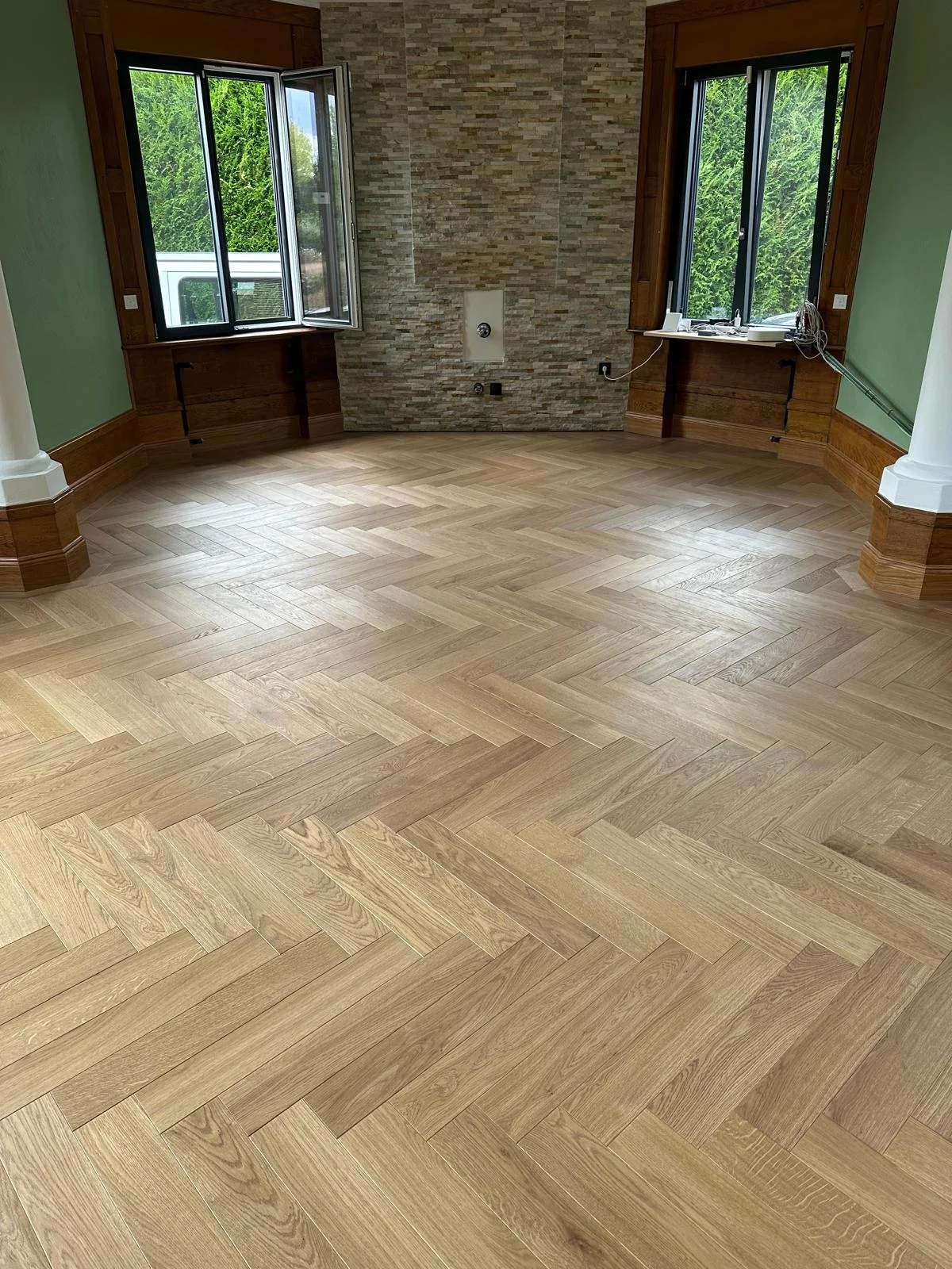 Pièce rénovée avec parquet en chêne à chevrons, murs en pierre et fenêtres ouvertes donnant sur une haie.