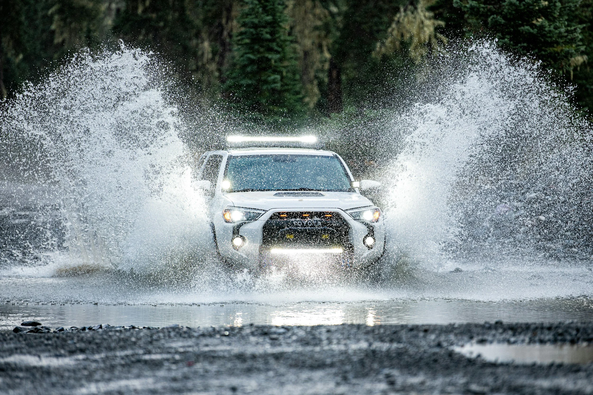 Ein weißes Offroadfahrzeug fährt durch einen Fluss, umgeben von einem bewaldeten Gebiet, bei Regen.