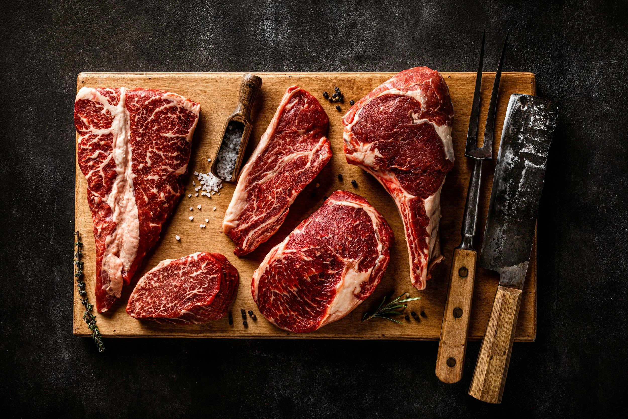 Verschiedene Stücke rotes Fleisch auf einem Holztisch mit Salz, Pfeffer und Küchenutensilien.