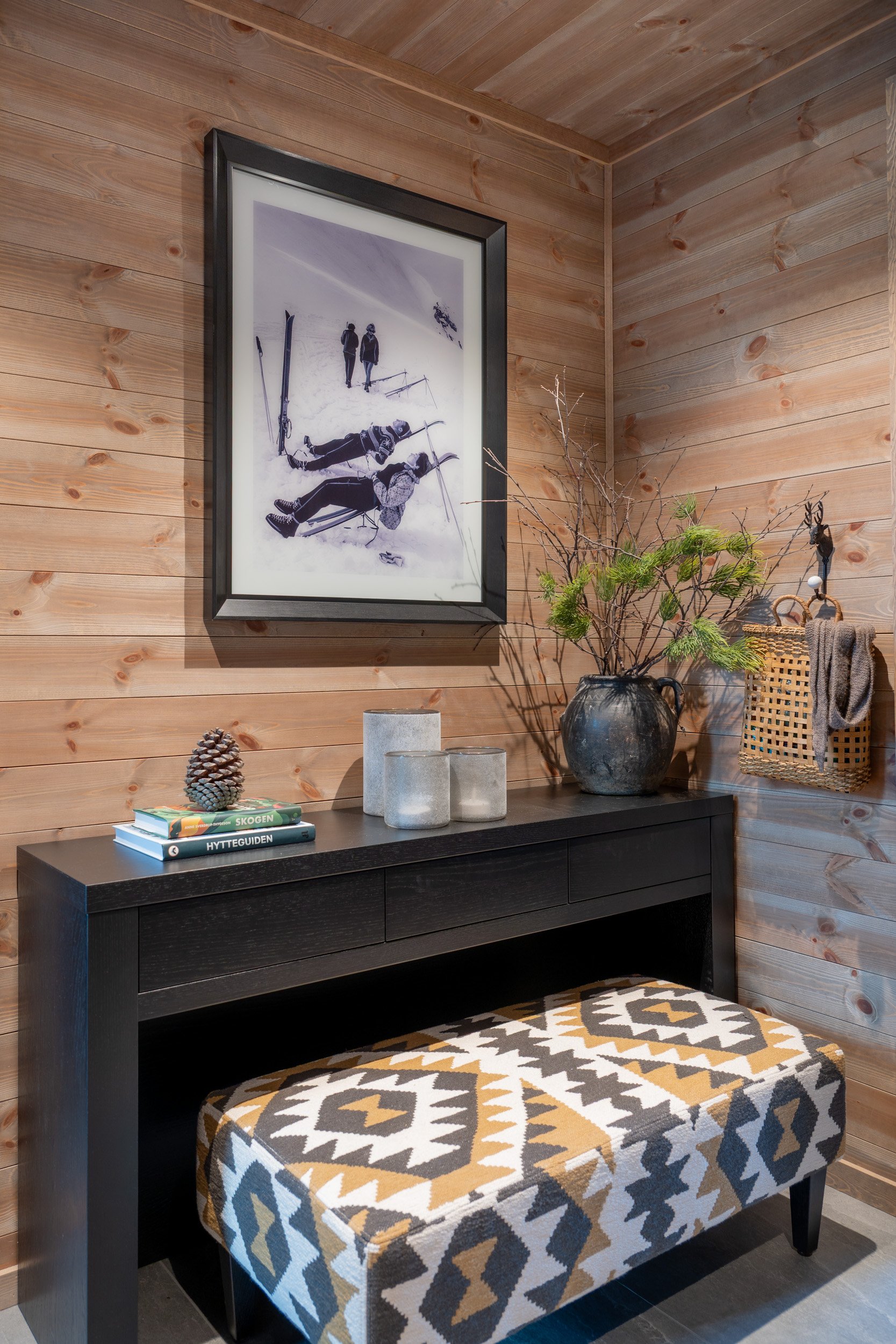 Innendørs hjørne med lyst trepanel, svart konsollbord med bøker, dekorativ pinecone, grå lykter, og en stor vase med kvister og en grønn plante. Under bordet en geometrisk mønstret pute.