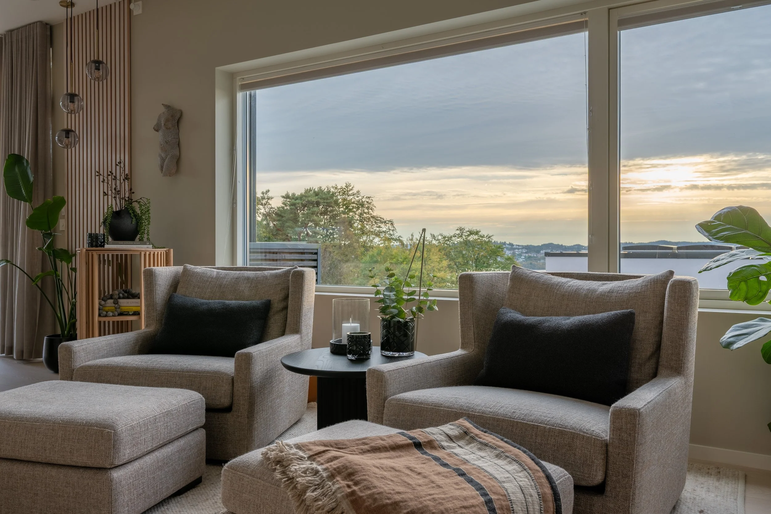 Stue med store vinduer som viser en utendørs utsikt med trær og solnedgang. Innendørs er det beige sofaer, grønne planter, dekorative lys, og et hjørne med en liten hylle.