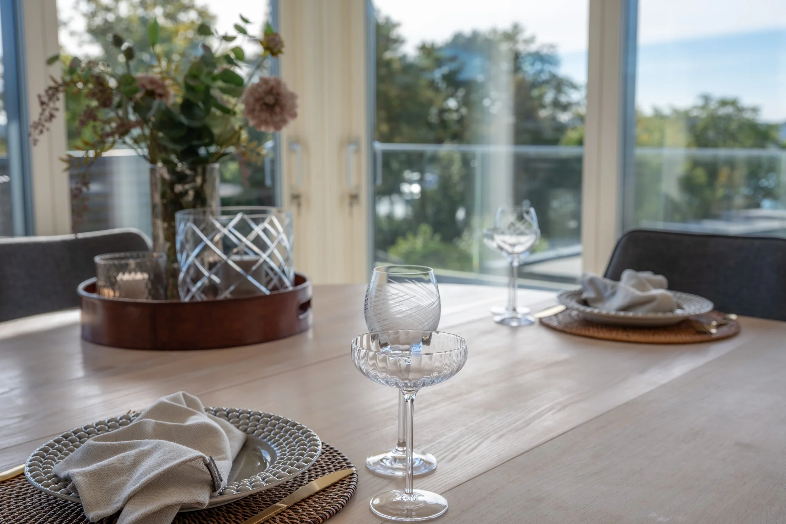 Bord med glass, tallerkener med servietter, en vase med blomster, og vindu med utsikt over trær utendørs.