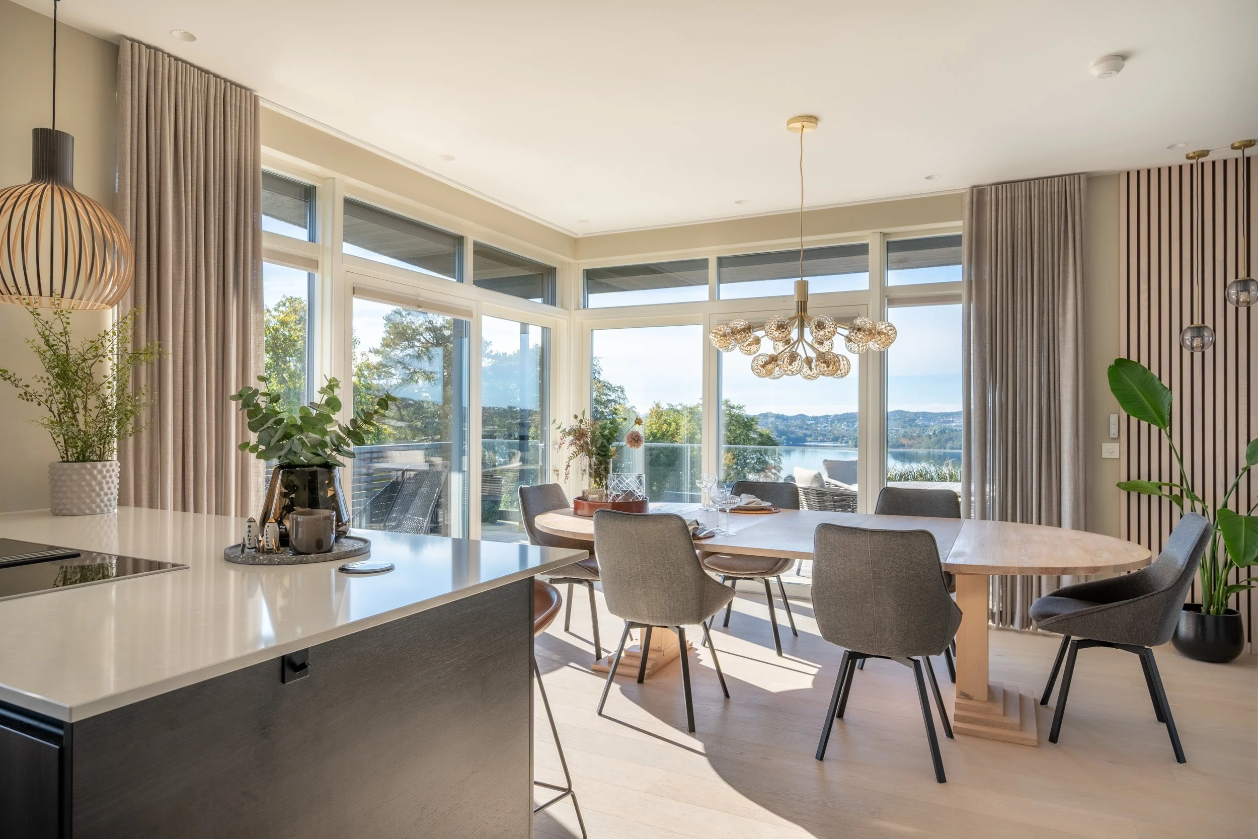 Innendørs kjøkken- og spiseområde med store vinduer som vender mot vann og natur. Det er et rundt spisebord med stolene rundt og blomsterdekorasjoner på bordet. Flere planter og moderne belysning henger fra taket.