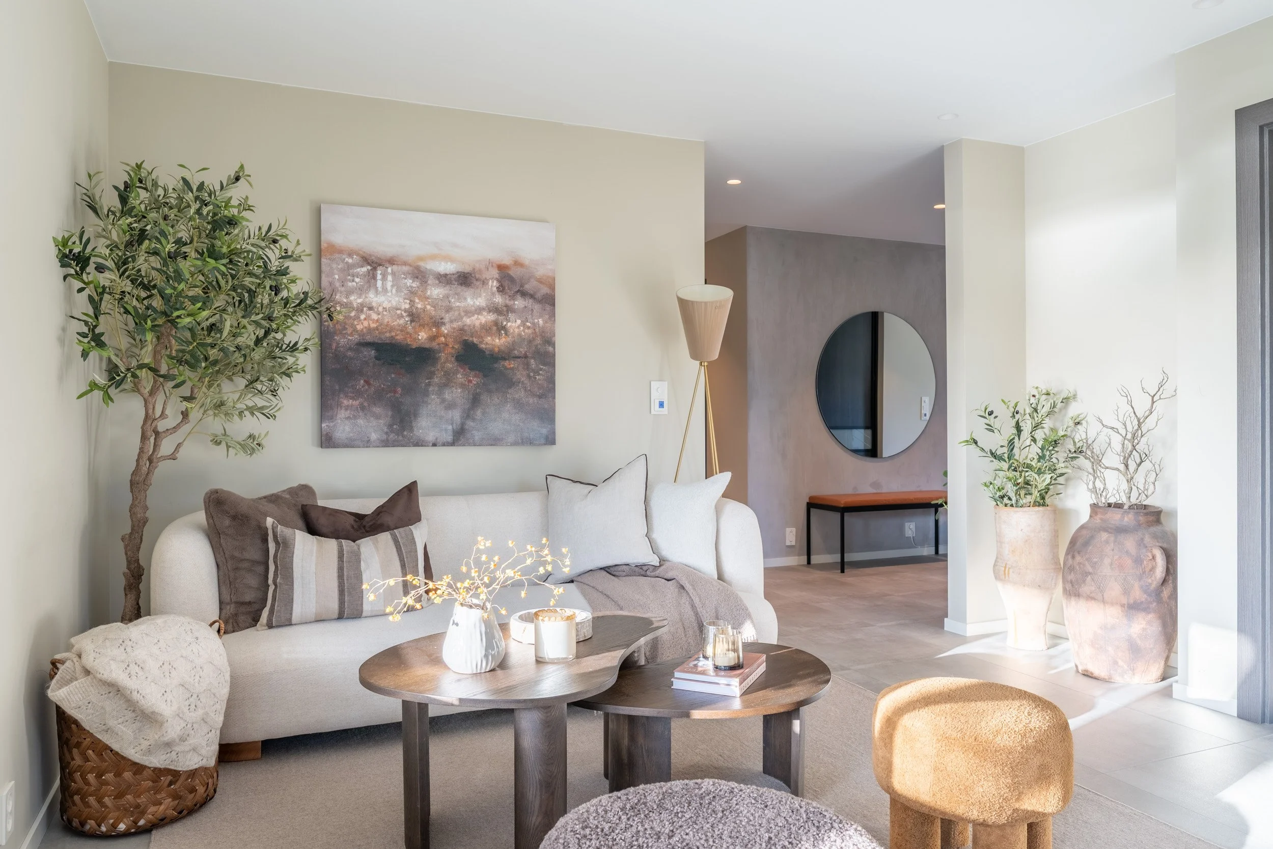 Innendørs stue med en hvit sofa, kunst på veggen, grønne planter, et rundt sofabord med dekorative gjenstander, og store keramikkvasker i hjørnet, alt i et lyst moderne hjem.
