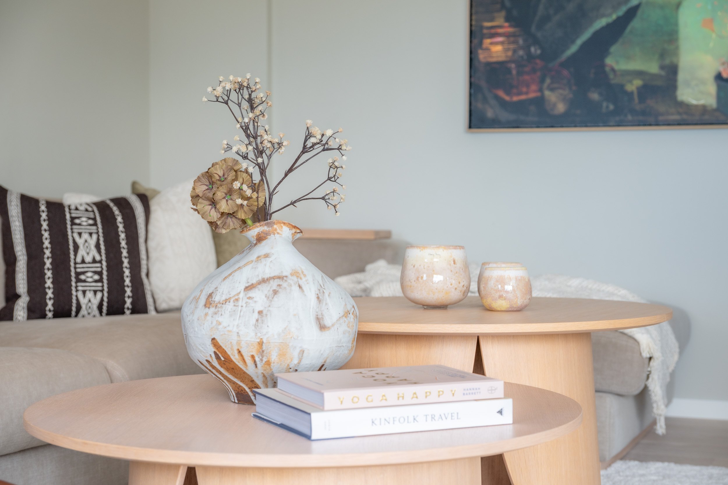 Innendørs scene med en dekorativ vase med tørkede blomster på et rundt trebord, og to mindre keramikkvaser ved siden av. I bakgrunnen en sofa med puter og et veggkunstverk.
