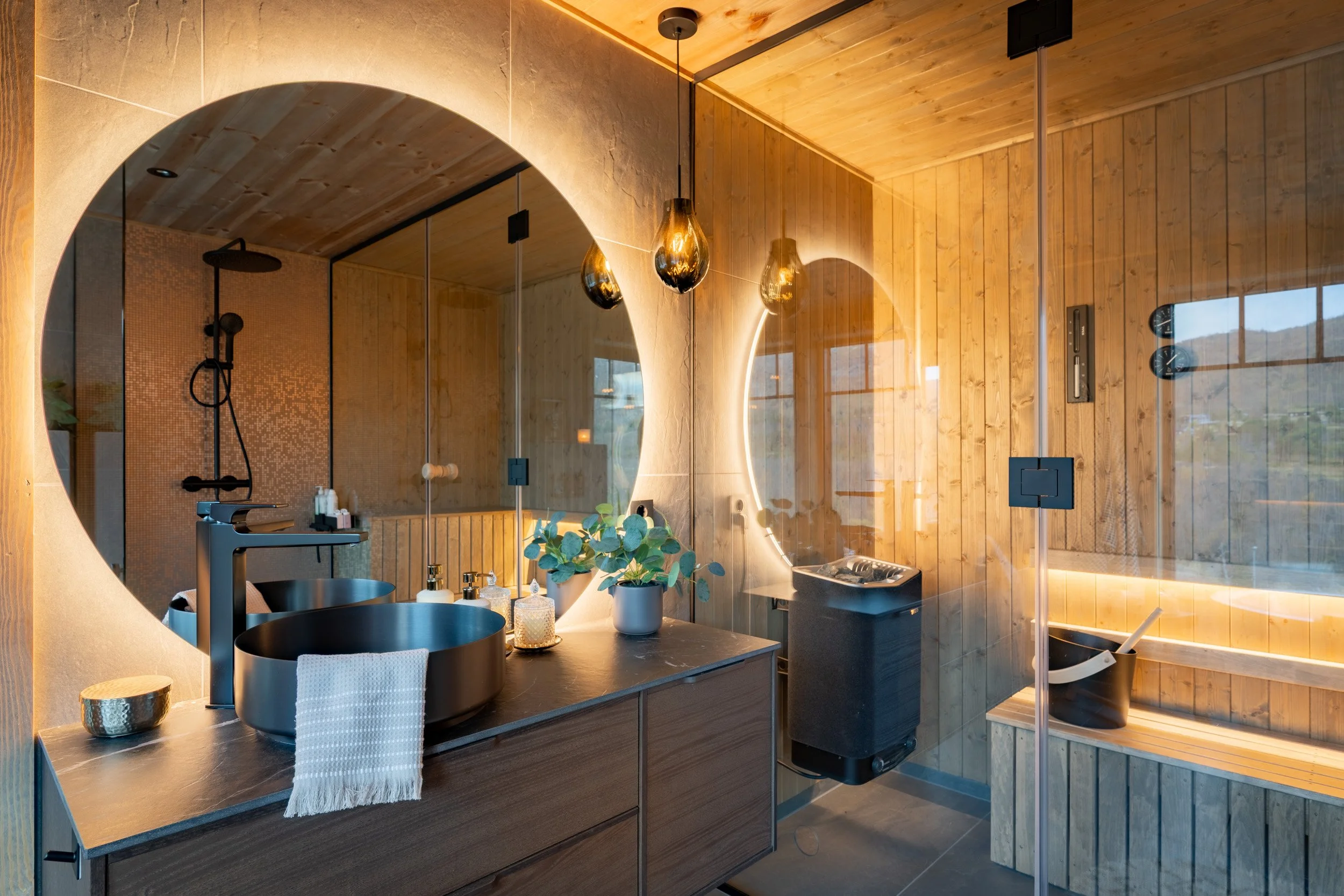 Et moderne bad med treramblede vegger og tak, glassdør til dusjrommet, håndvask med svart servant, speil med LED-belysning, hengende pendellamper, og grønne planter på vaskebenken.