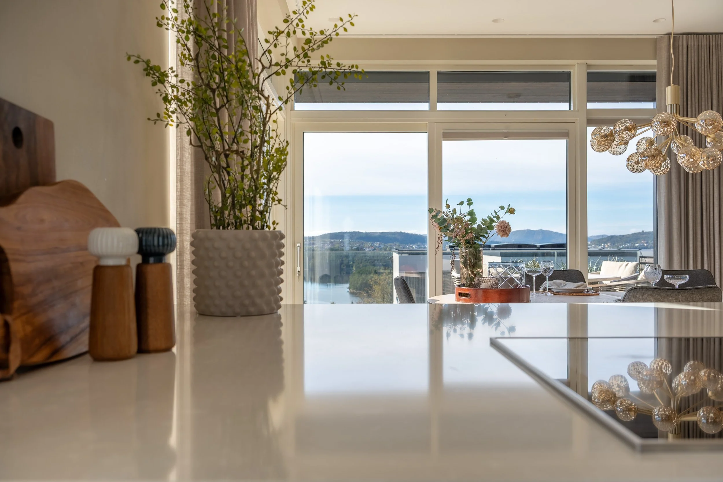 Innendørs bilde av et rom med store vinduer, planter i hvite og mørke potter, og et spisebord med glass, tallerkener og bestikk. Utsikt over landskap med fjell og vann i bakgrunnen.