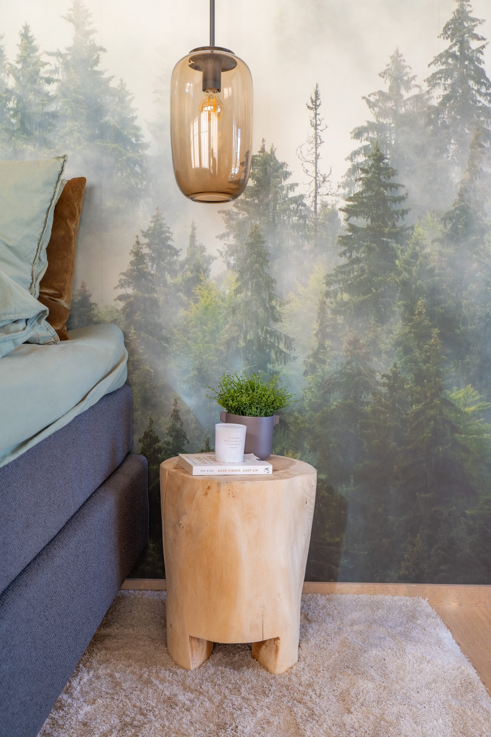 Et lite rundt trestykke brukt som bord med en bok og et lite lys på, plassert på en beige vegg med bilde av tette grantrær som bakgrunn, i en stue med en sofa og tepper.