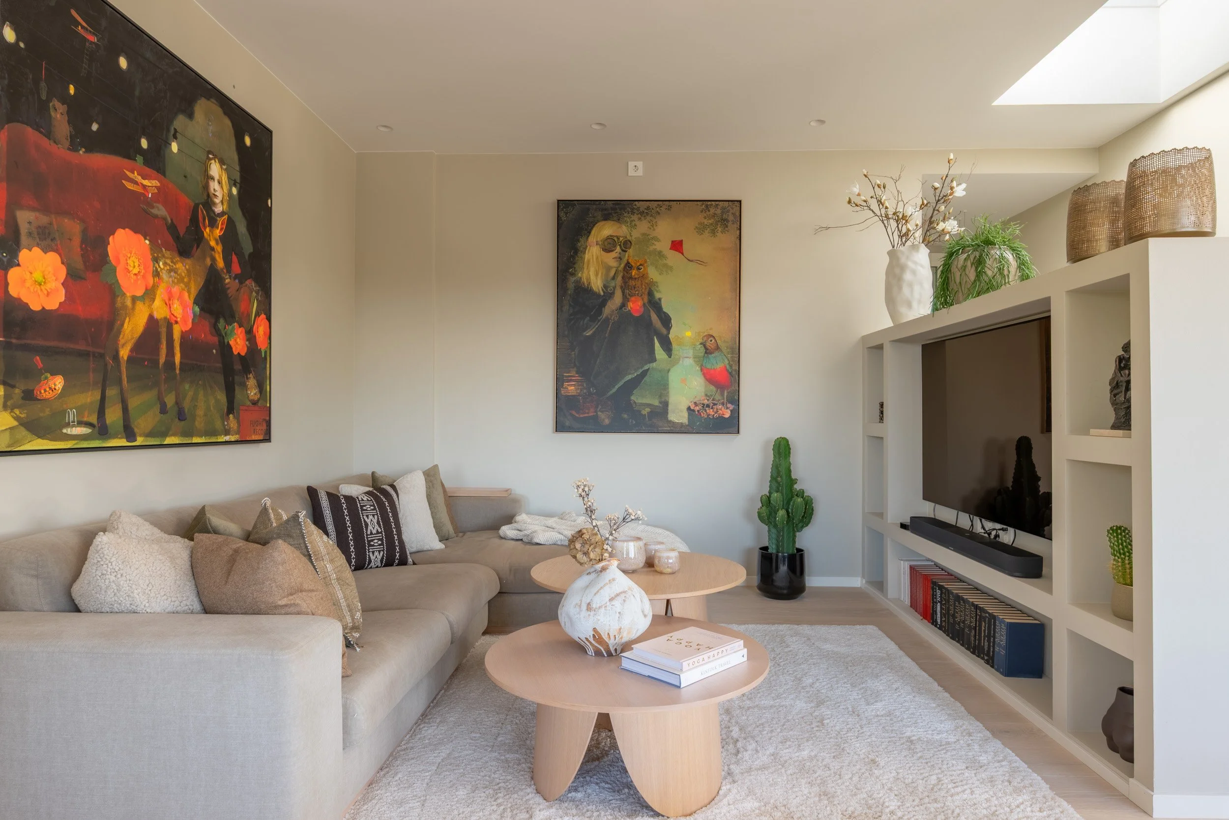 Et moderne stueinnredning med beige sofa, løse puter, små rundt trebord, kunstverk på veggen, grønne planter, et TV-skap, og et vindu i taket som slipper inn dagslys.