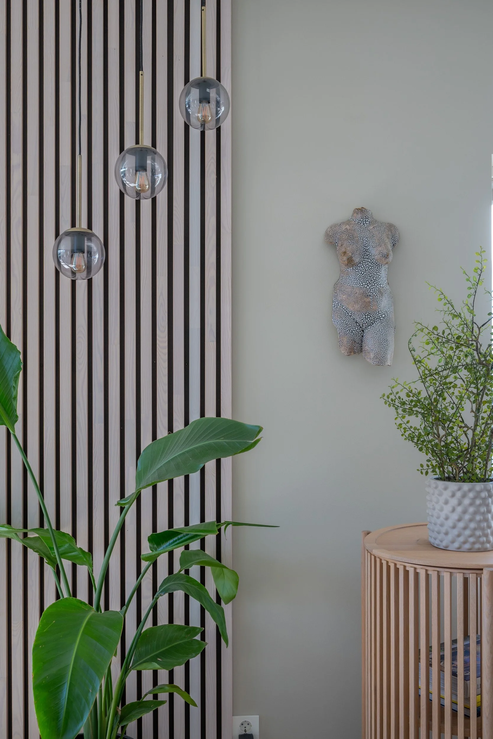 Et interiør med tre lameller på veggen, tre glasskulependler og en kunstnerisk kvinneliknende skulptur på veggen, omgitt av grønne planter.