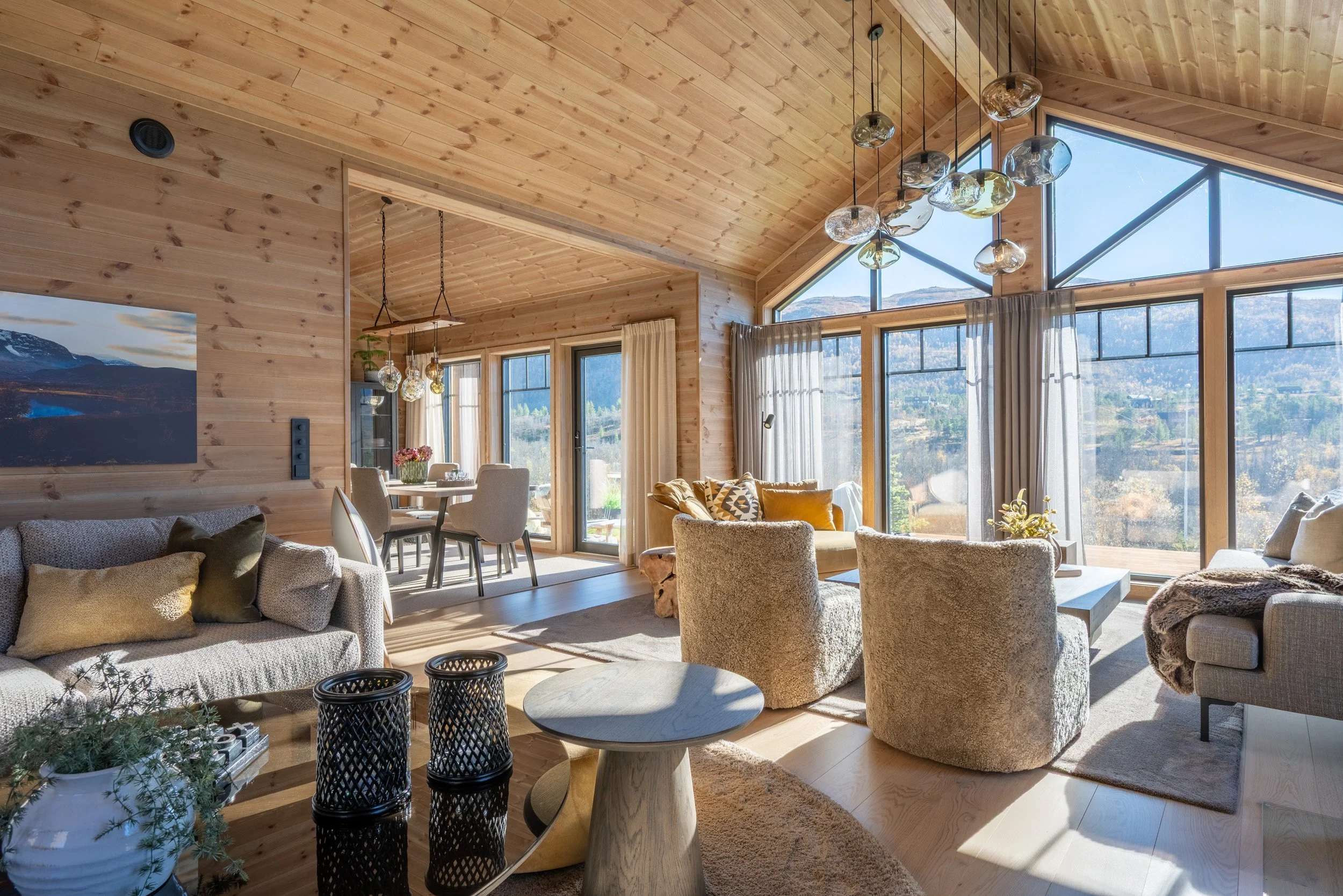 Innendørs stue med store vinduer, trebjelkeloft og moderne belysning, stuemøbler som sofa og stoler.