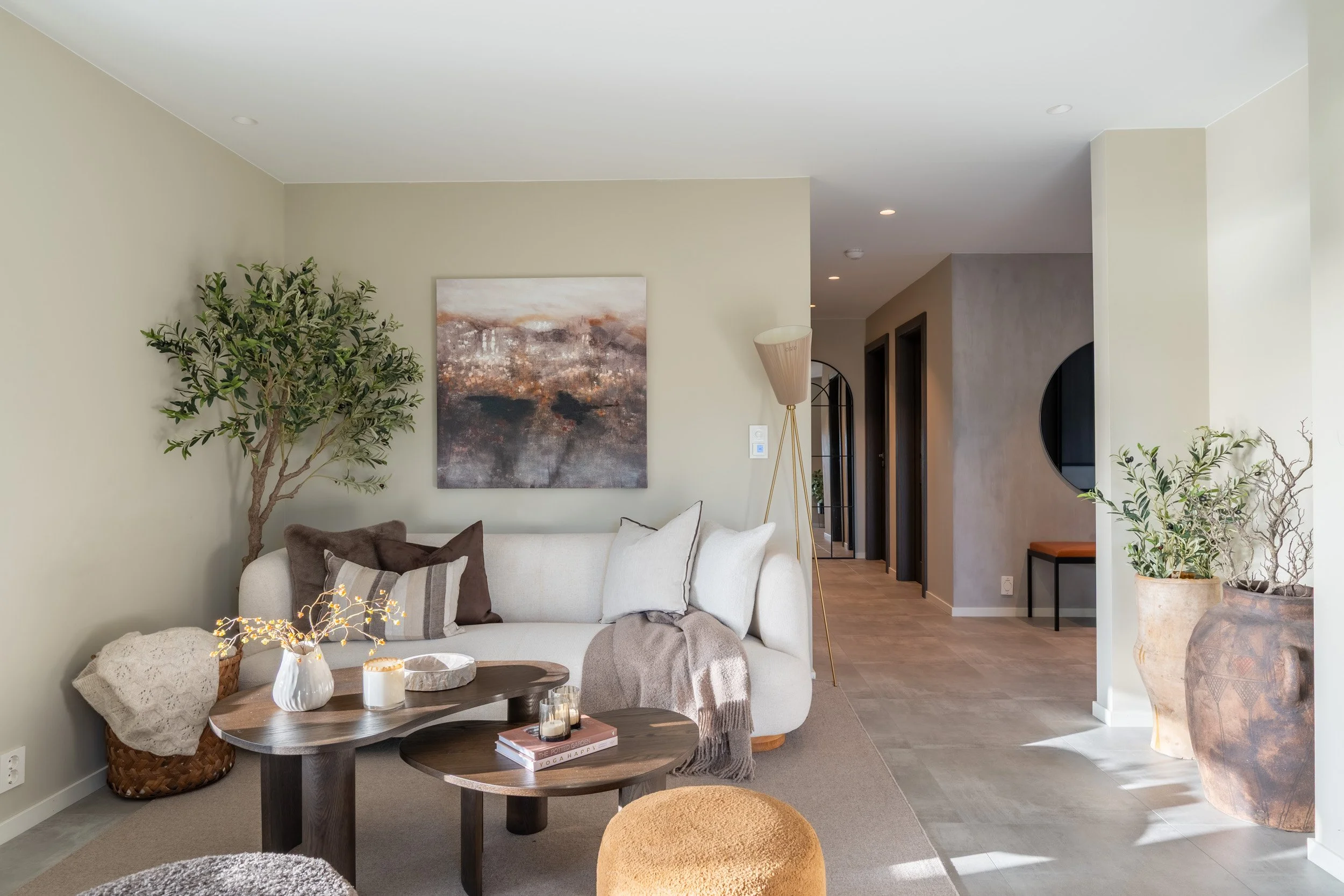 Stue med beige sofa, rundt barbord med lysestaker, kunstverk på veggen, planter, gul gulvlampe, og et fellesrom med dører og kurvfottør.