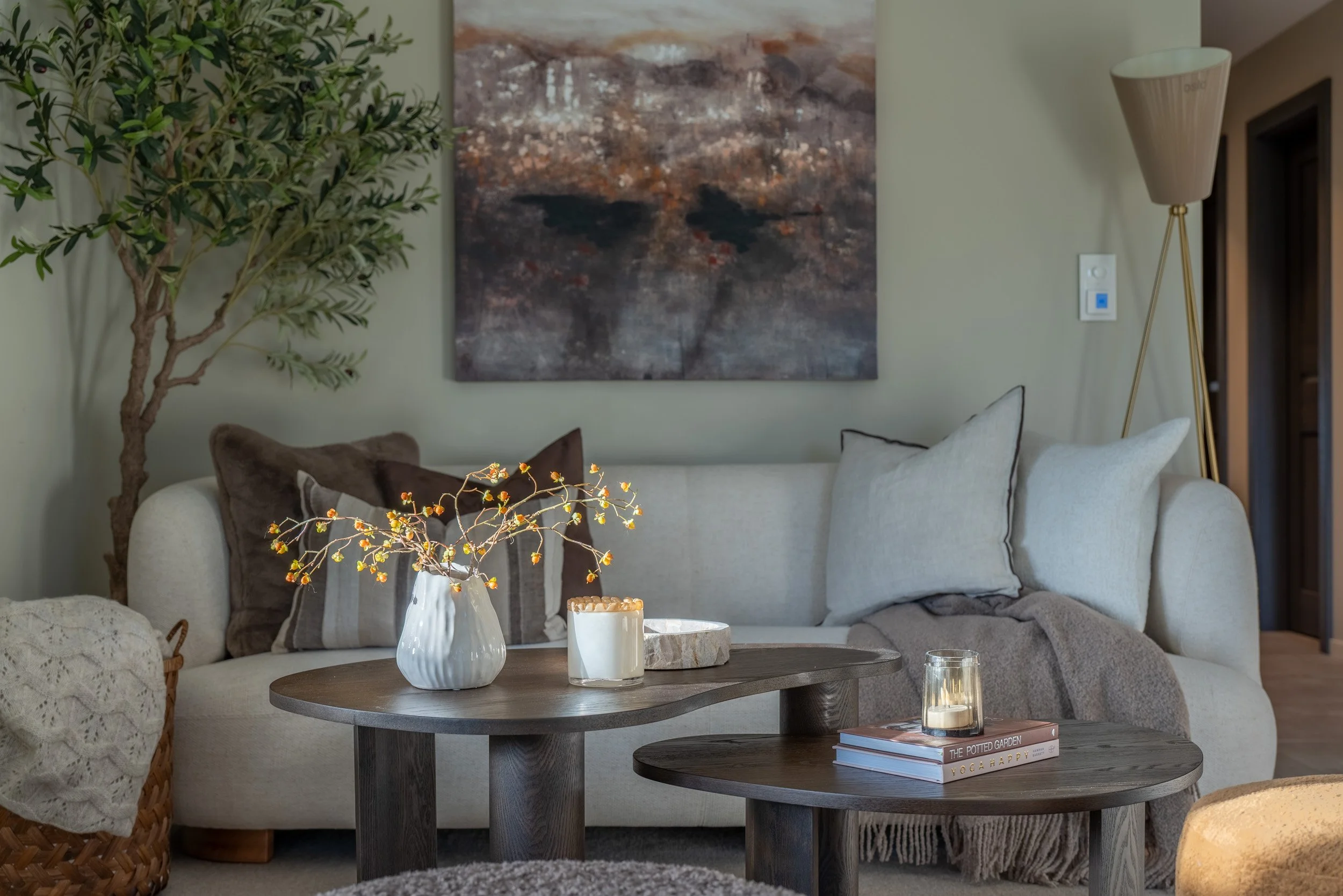 Stue med sofaputer, to runde trekongrepet bord med høye lys, bladkledd plante, stort maleri på veggen, bøker, lyslykt og krydder på bordet.