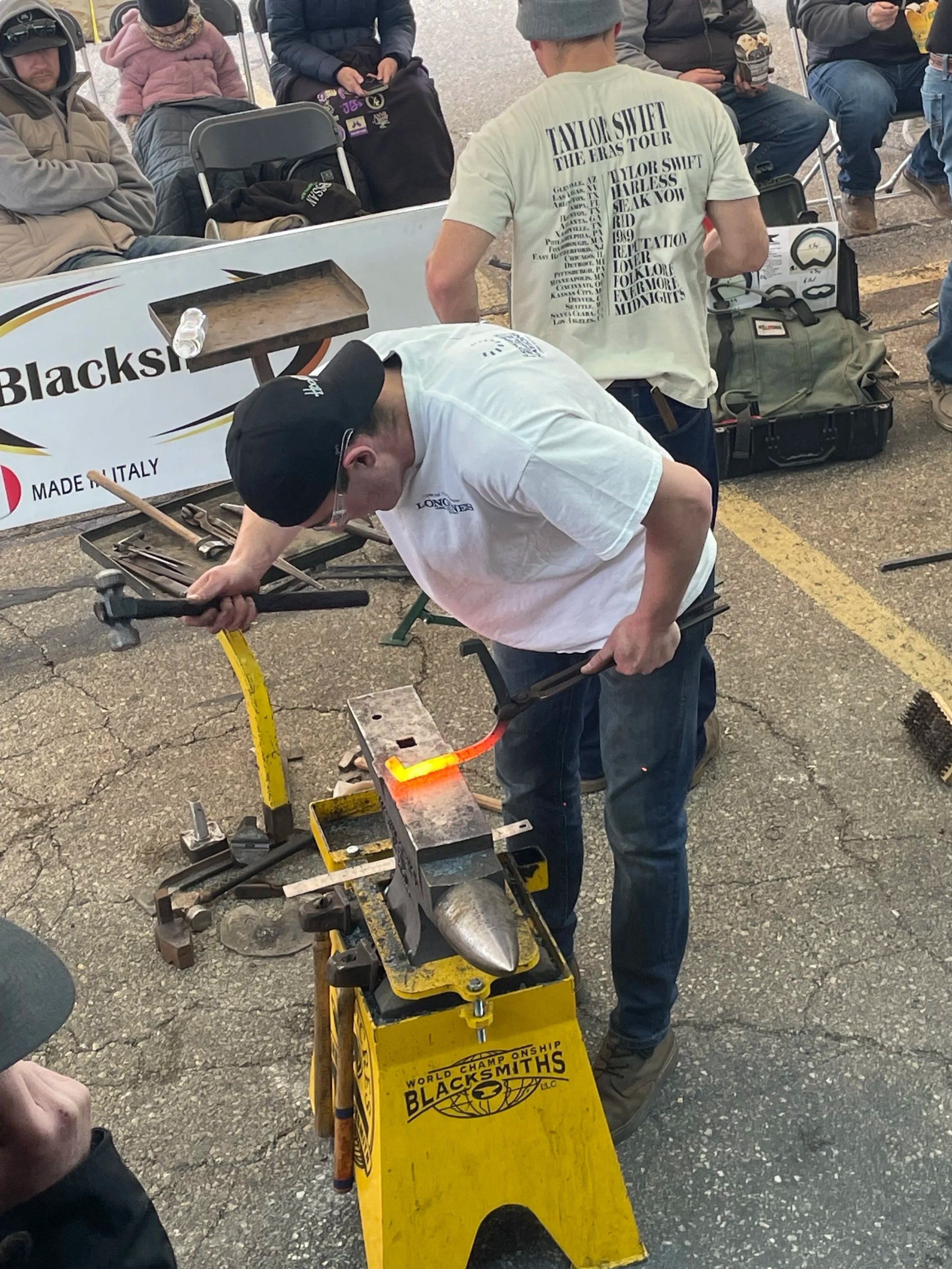 A man working on a blacksmith project with various tools, surrounded by spectators at an outdoor event. The blacksmith's cart has 'World Championship Blacksmiths' printed on it.