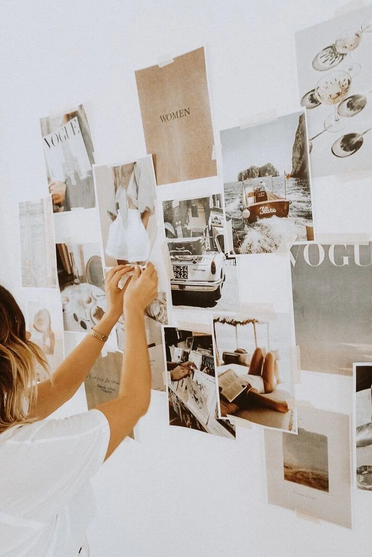 Person pinning photographs and magazine cutouts on a white wall, including images of cars, a boat, a person reading, and fashion-related images.