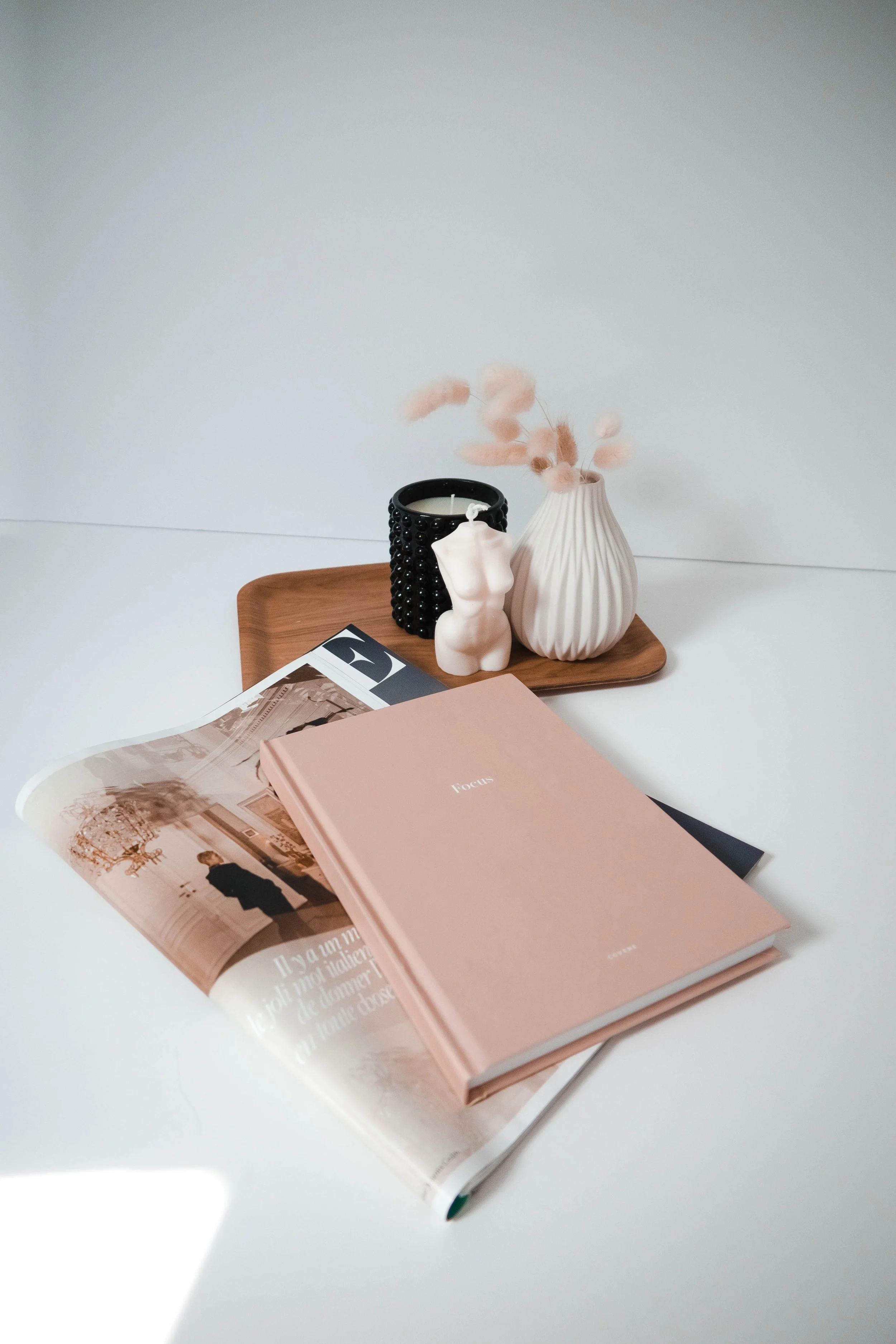 A wooden tray on a white surface holds a white vase with pink dried flowers, a black textured candle holder with a white candle, a small white torso statue, and magazines with pastel covers, including a pink one labeled 'Focus'.