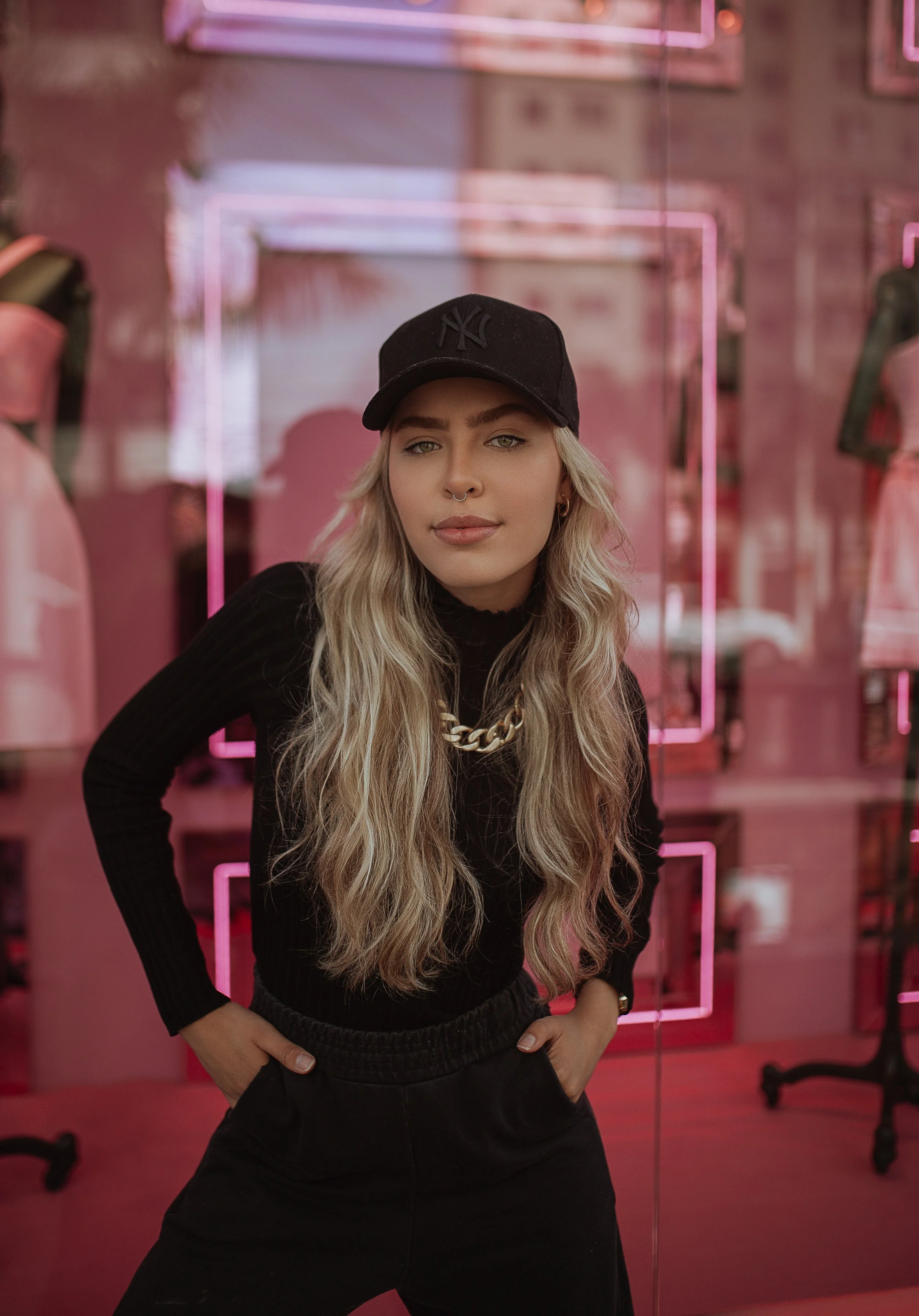 A young woman with long blonde hair, wearing a black New York Yankees cap, black long-sleeve top, gold chain necklace, and yellow contact lenses, posing with her hands on her hips in front of a pink neon-lit mirror.