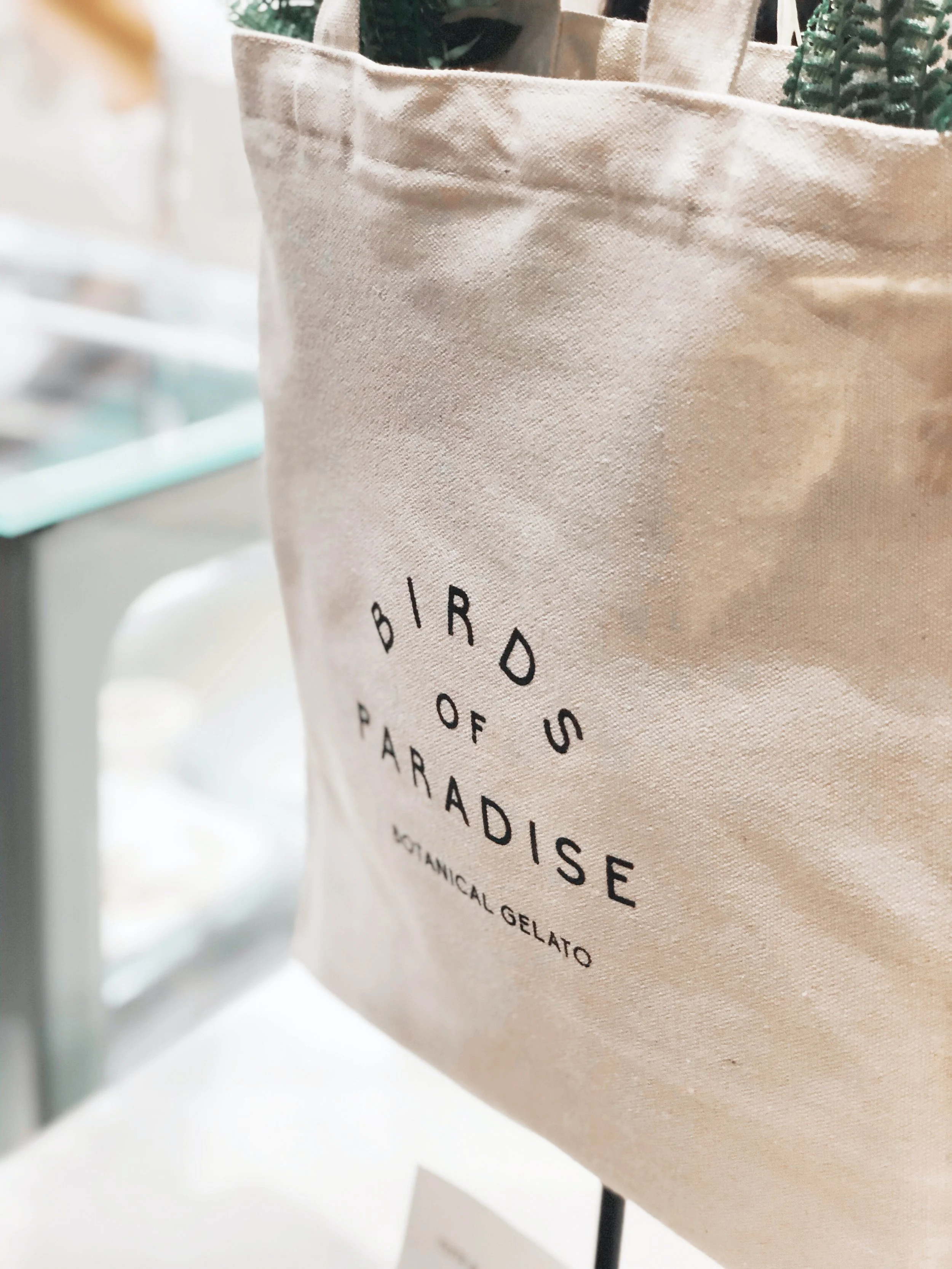 Beige tote bag with black text reading "BIRD OF PARADISE, WOODLAND GELATO" and some green plants inside.