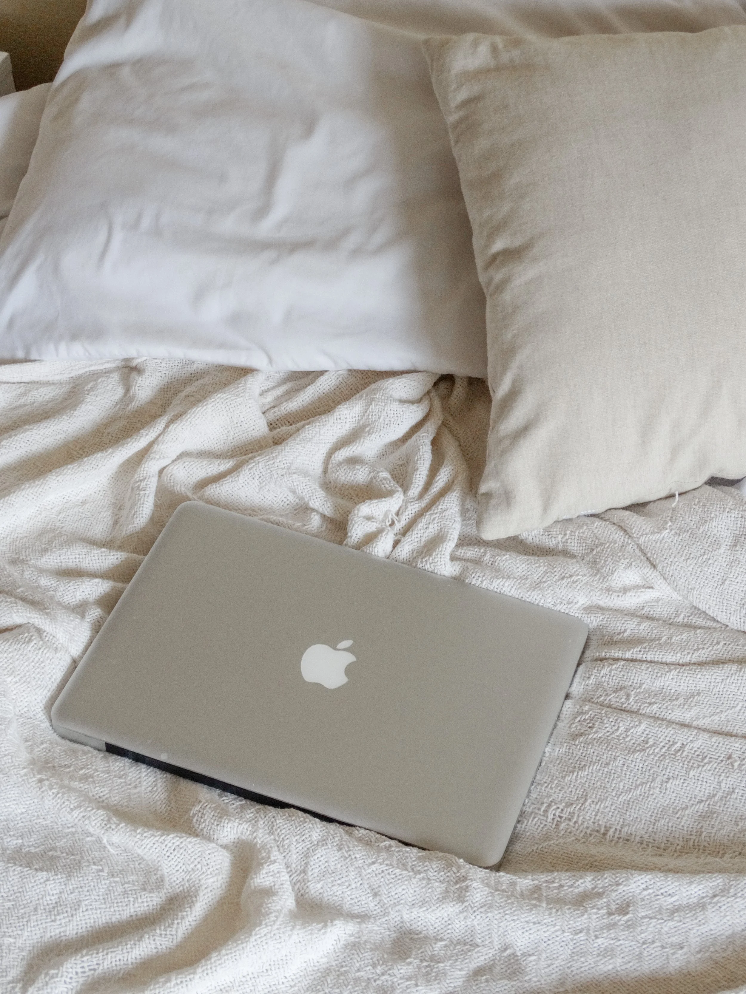 A closed silver MacBook laptop on a bed with white and cream-colored pillows and a textured blanket.