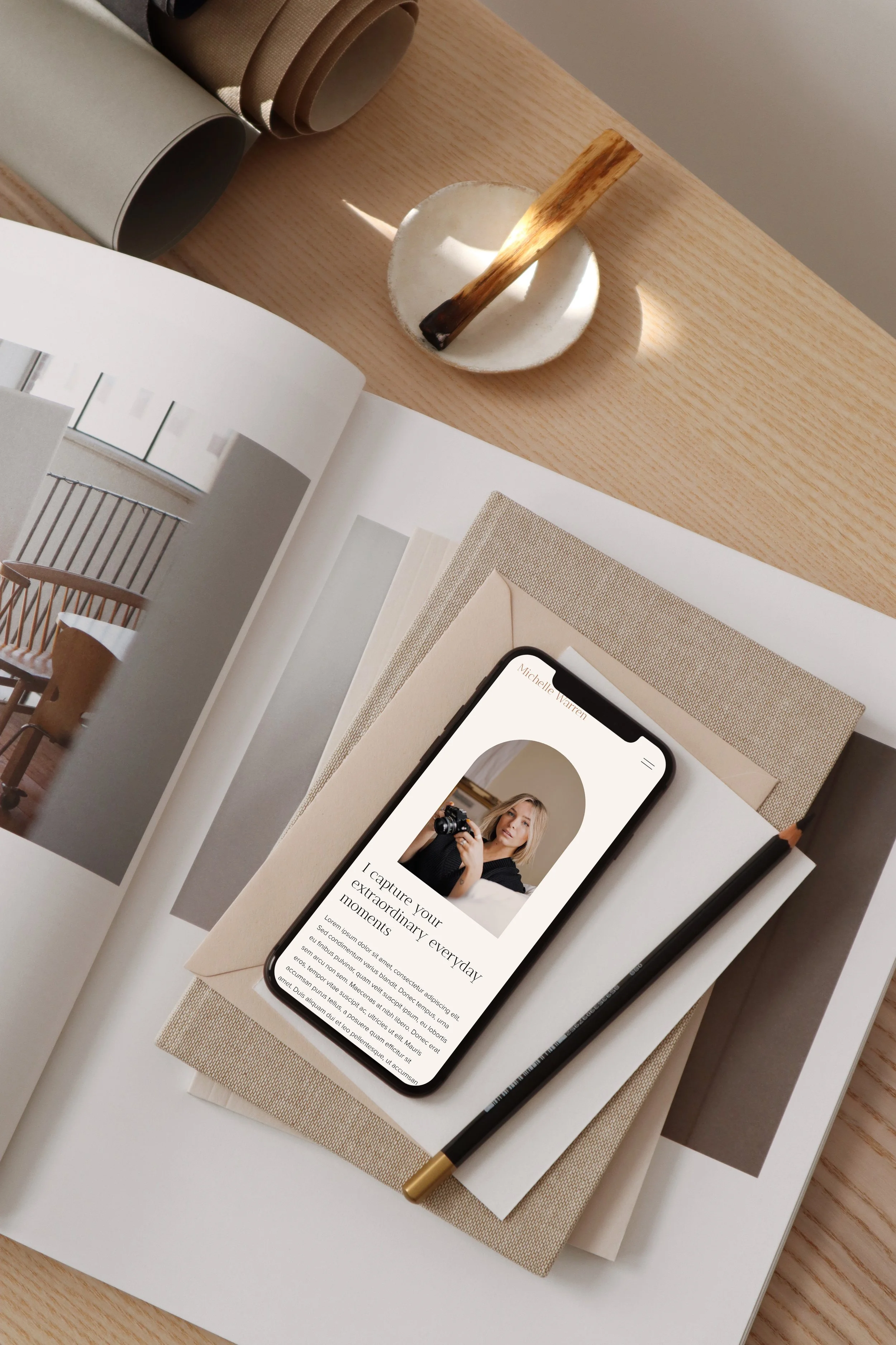A flat lay of a wooden table with a magazine, a smartphone displaying a woman with a camera, a beige notebook, and a black pen. There are also three rolled-up pieces of fabric and a small white dish with a wooden spatula.