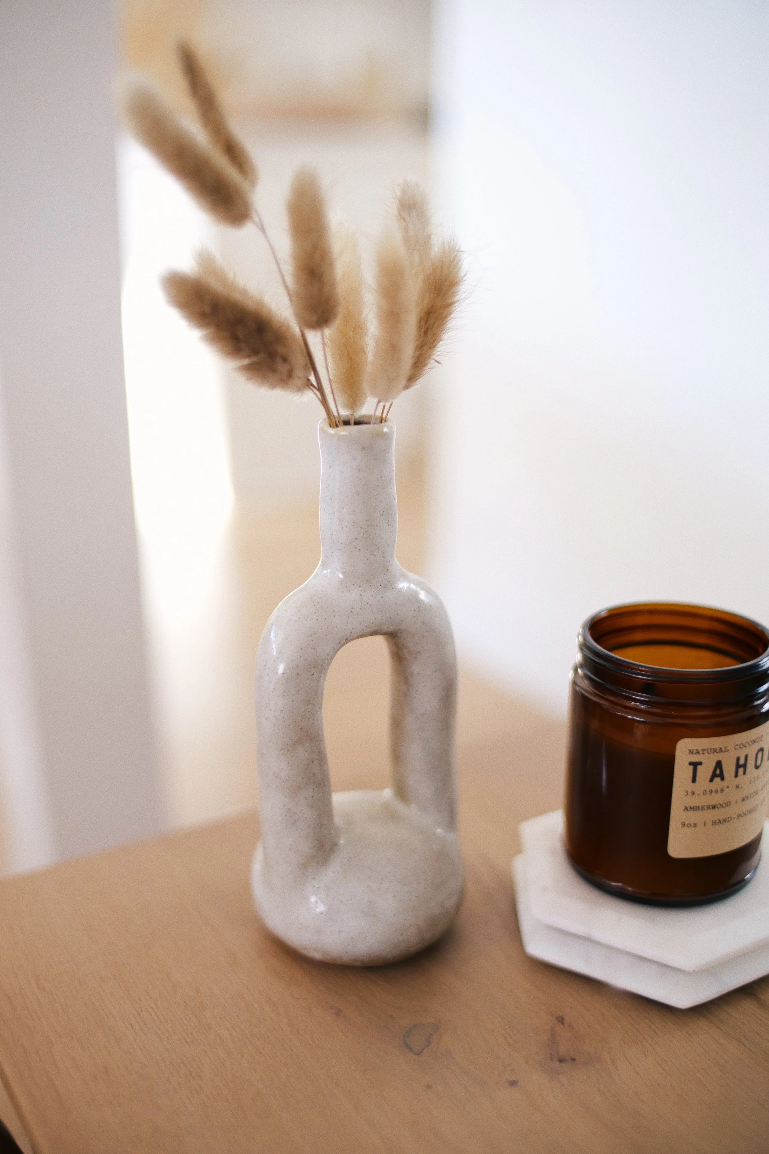 A white ceramic vase with an artistic, modern design holds dried beige and light brown grasses. On the right, there is a dark amber glass jar with a white label, placed on a small white tray on a wooden surface.