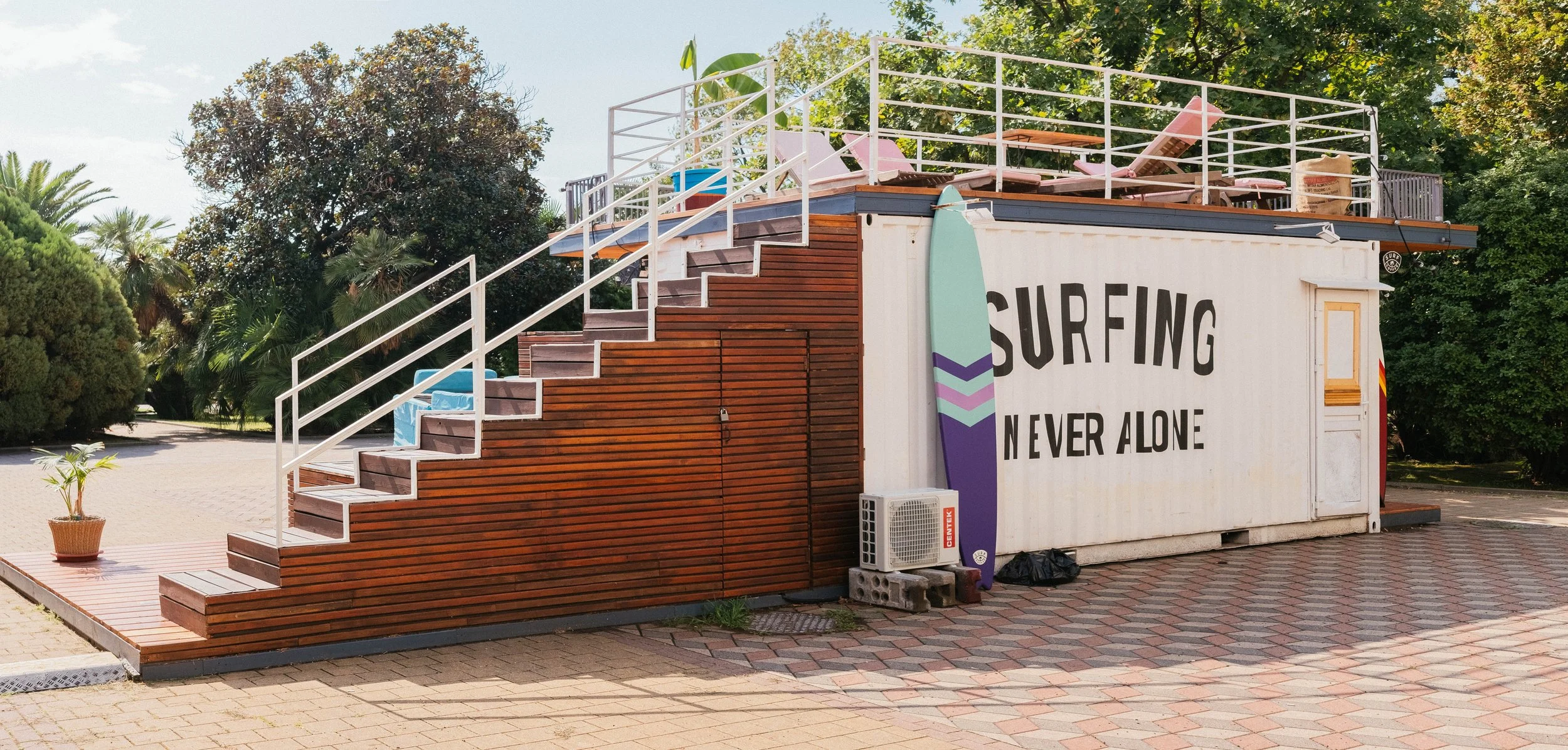 A small building with a sign that reads 'Surfing Never Alone' and a colorful surfboard graphic. The building has wooden stairs leading to the rooftop area equipped with lounge chairs and surrounded by trees and greenery.