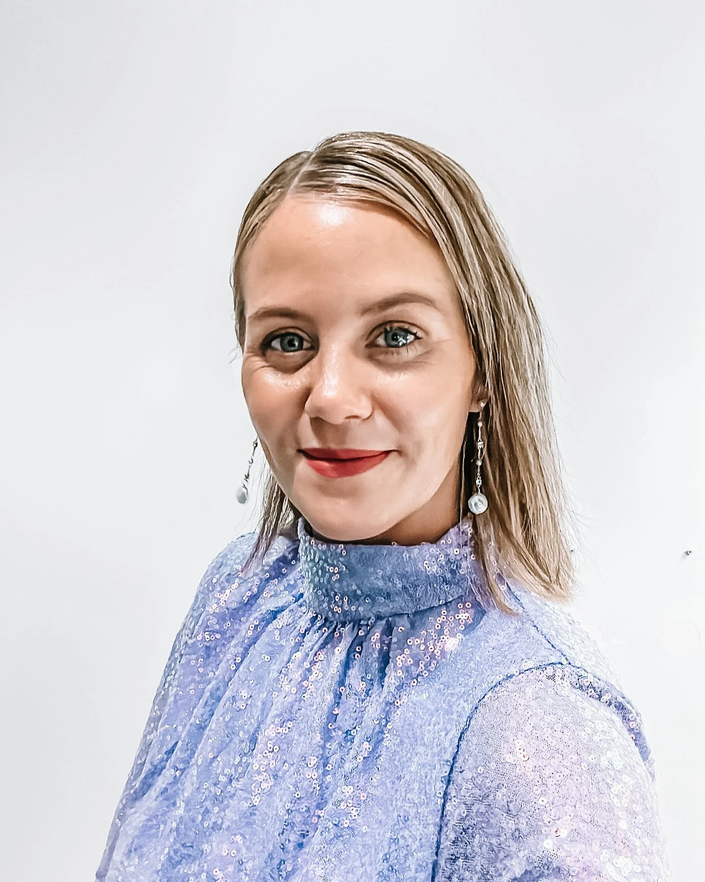A woman with blonde hair and blue eyes smiling at the camera, wearing a light purple, sequined top and earrings, against a plain white background.