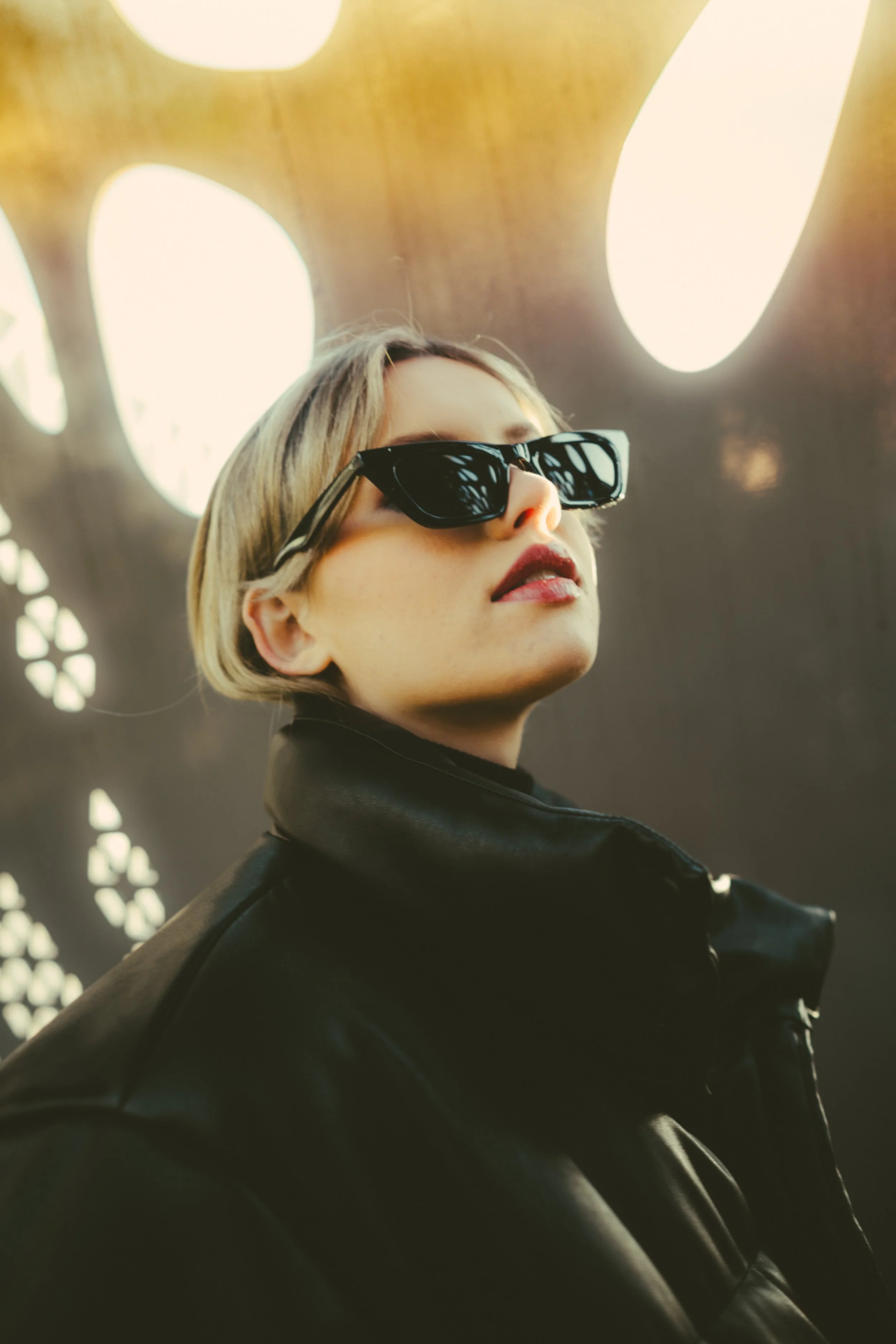 A woman with short blonde hair wearing black sunglasses and a black leather jacket against a background with large glowing lights.