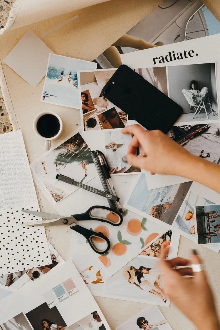 A cluttered desk with photos, scissors, a black marker, a pair of hands, a coffee cup, a smartphone, and various printed materials.