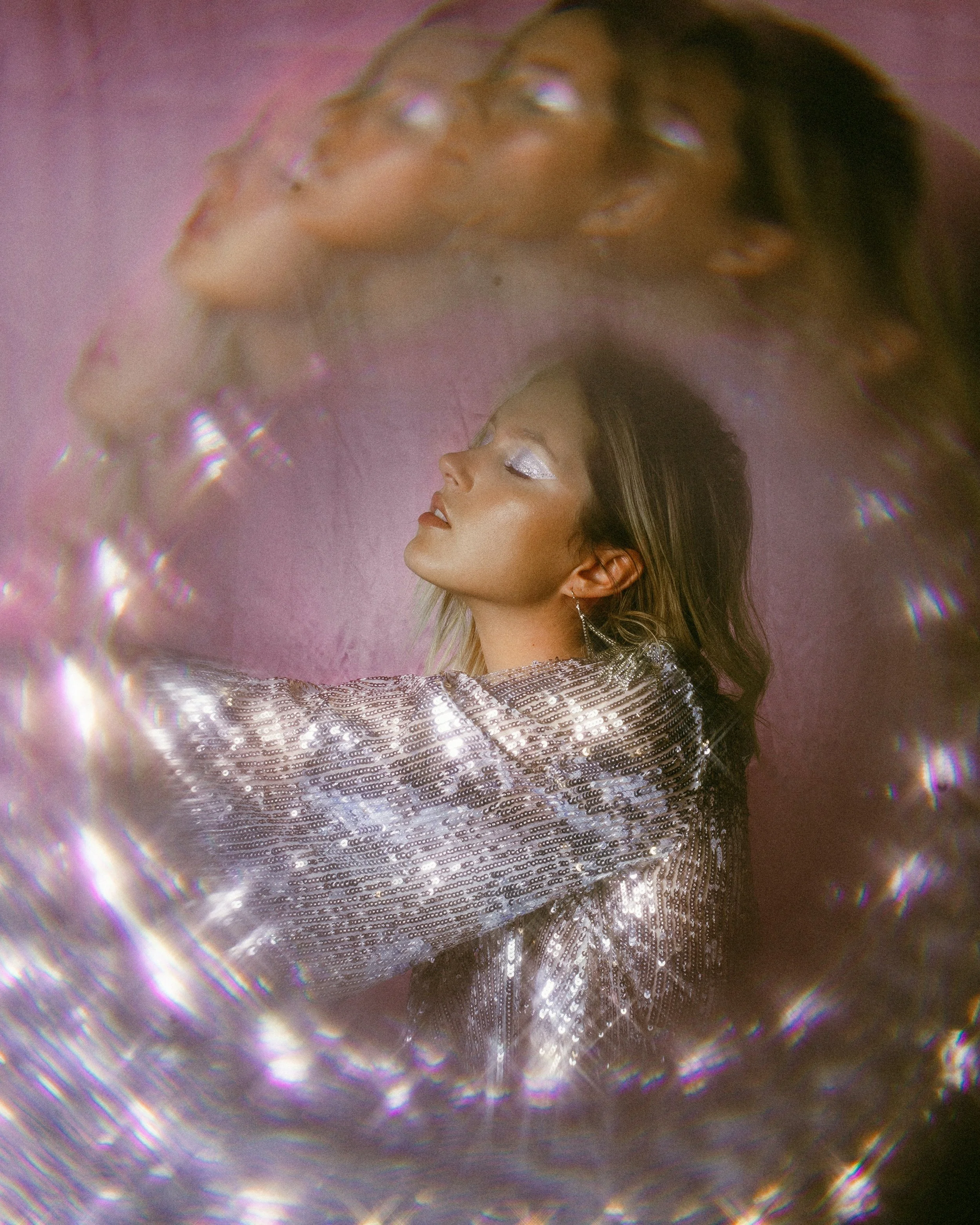 A woman with silver makeup and earrings, dressed in a shiny, metallic outfit, posed against a pink background with a reflection or distortion effect.