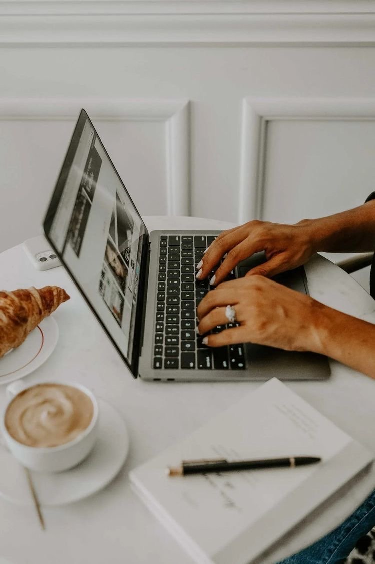 Person typing on a laptop at a white table with a notebook and pen, a croissant, and a cup of coffee, against a white paneled wall.