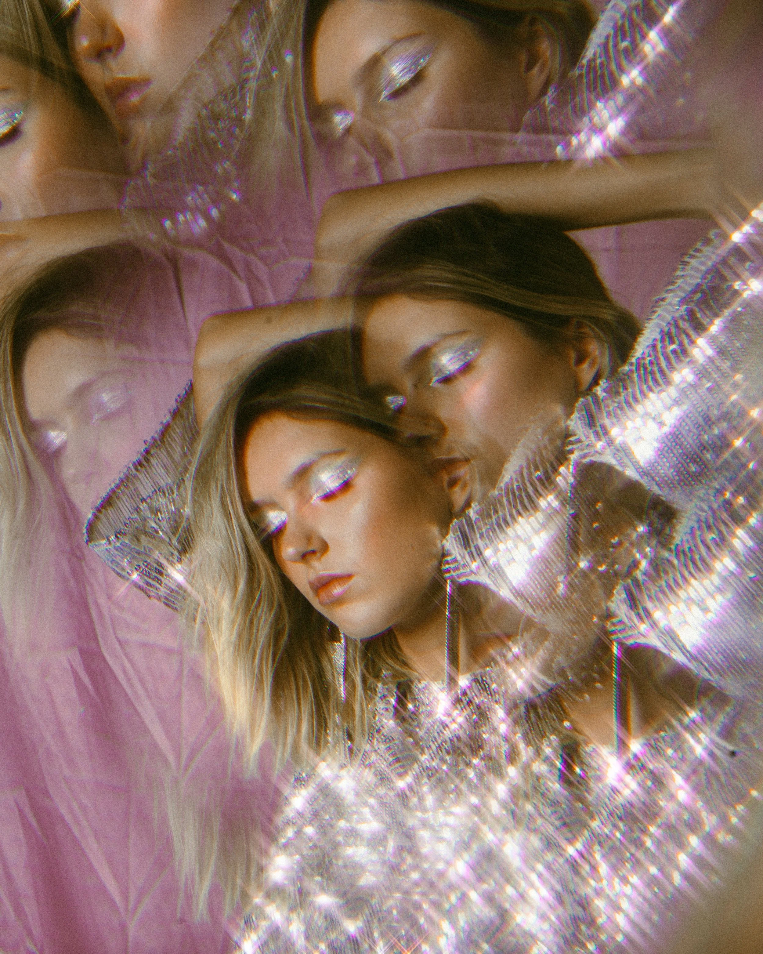 Close-up of a young woman with long brown hair, wearing a pink outfit and glittery eyeshadow, lying on a pink surface, with a fragmented mirror reflecting her face and the surrounding scene.