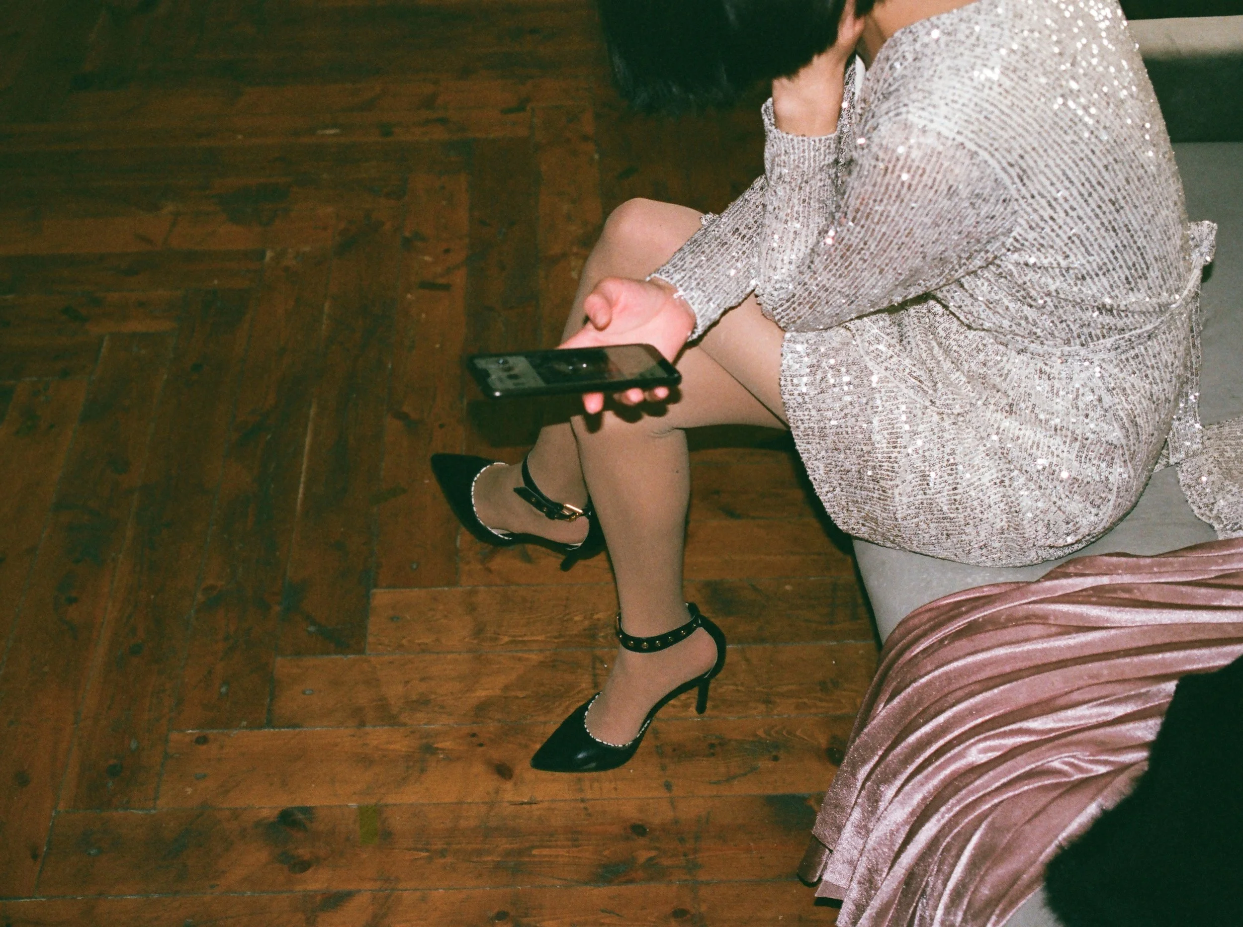 Woman sitting on a bench wearing a sparkly, sequined dress and black high heels, holding a smartphone.