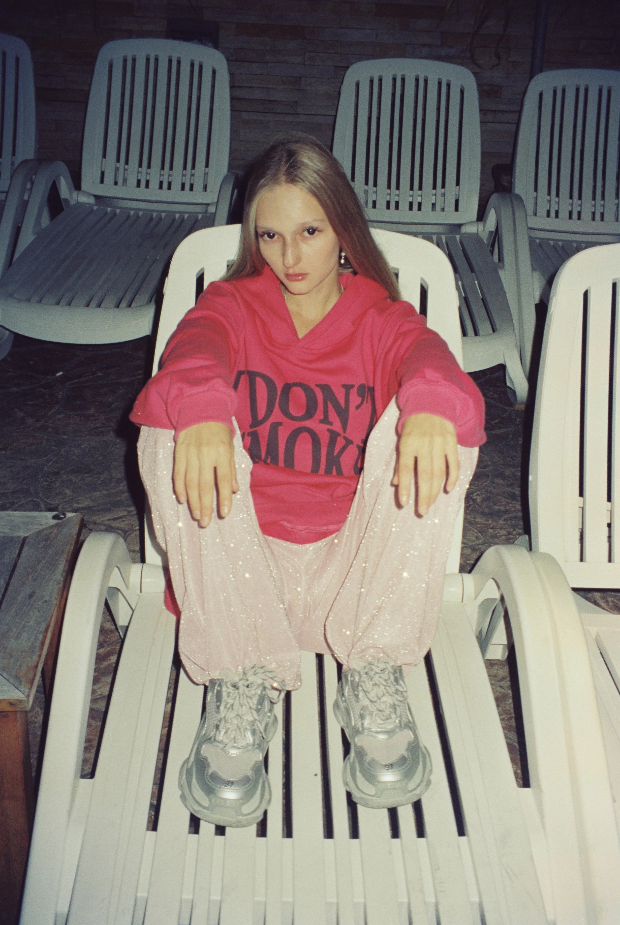 A young woman in a red hoodie and sparkly pink pants sitting on a white plastic lounge chair in an outdoor setting, with several empty similar chairs in the background against a brick wall.