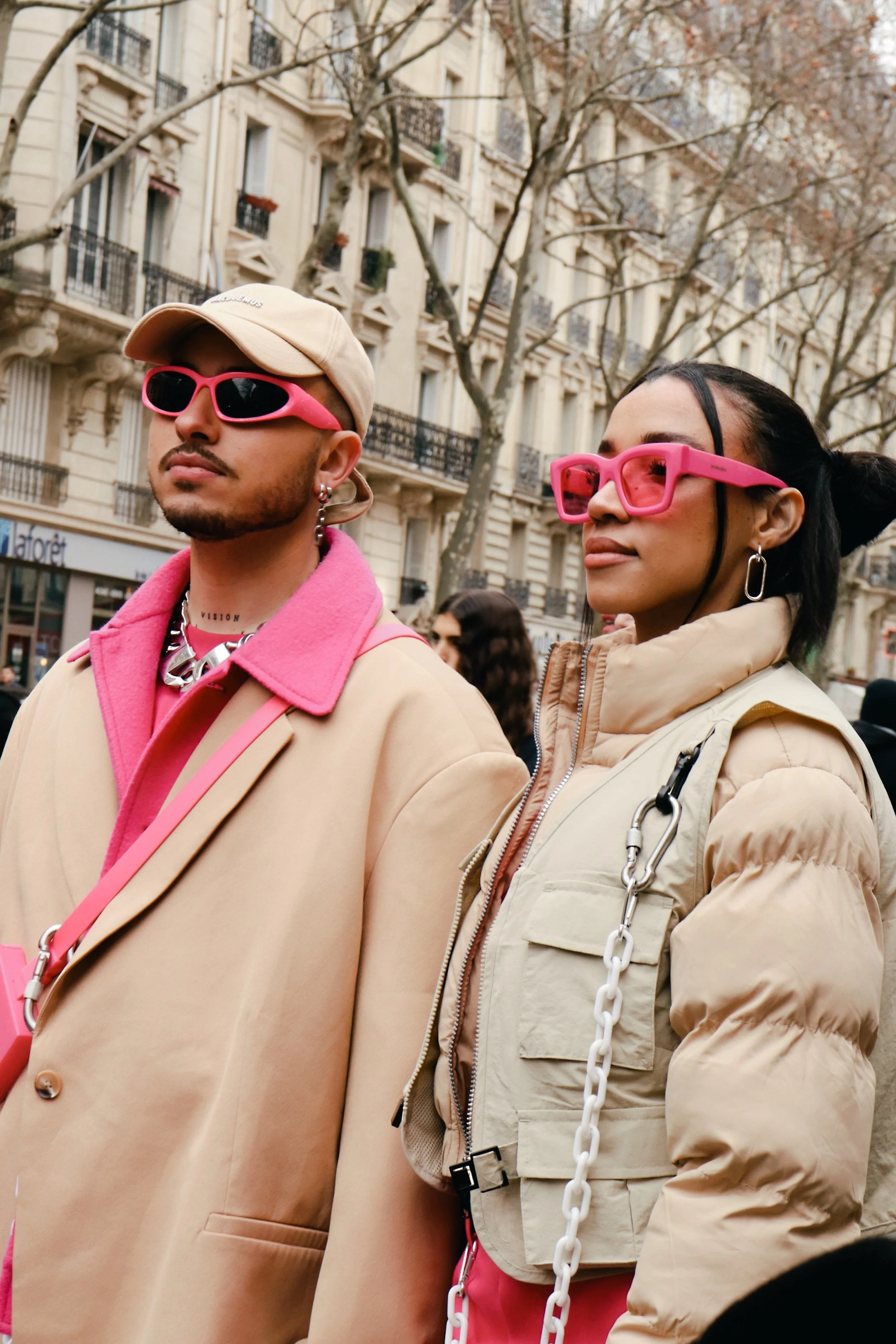 Two individuals standing outdoors in front of a building, wearing beige clothing and bright pink sunglasses, with trees and a cityscape in the background.