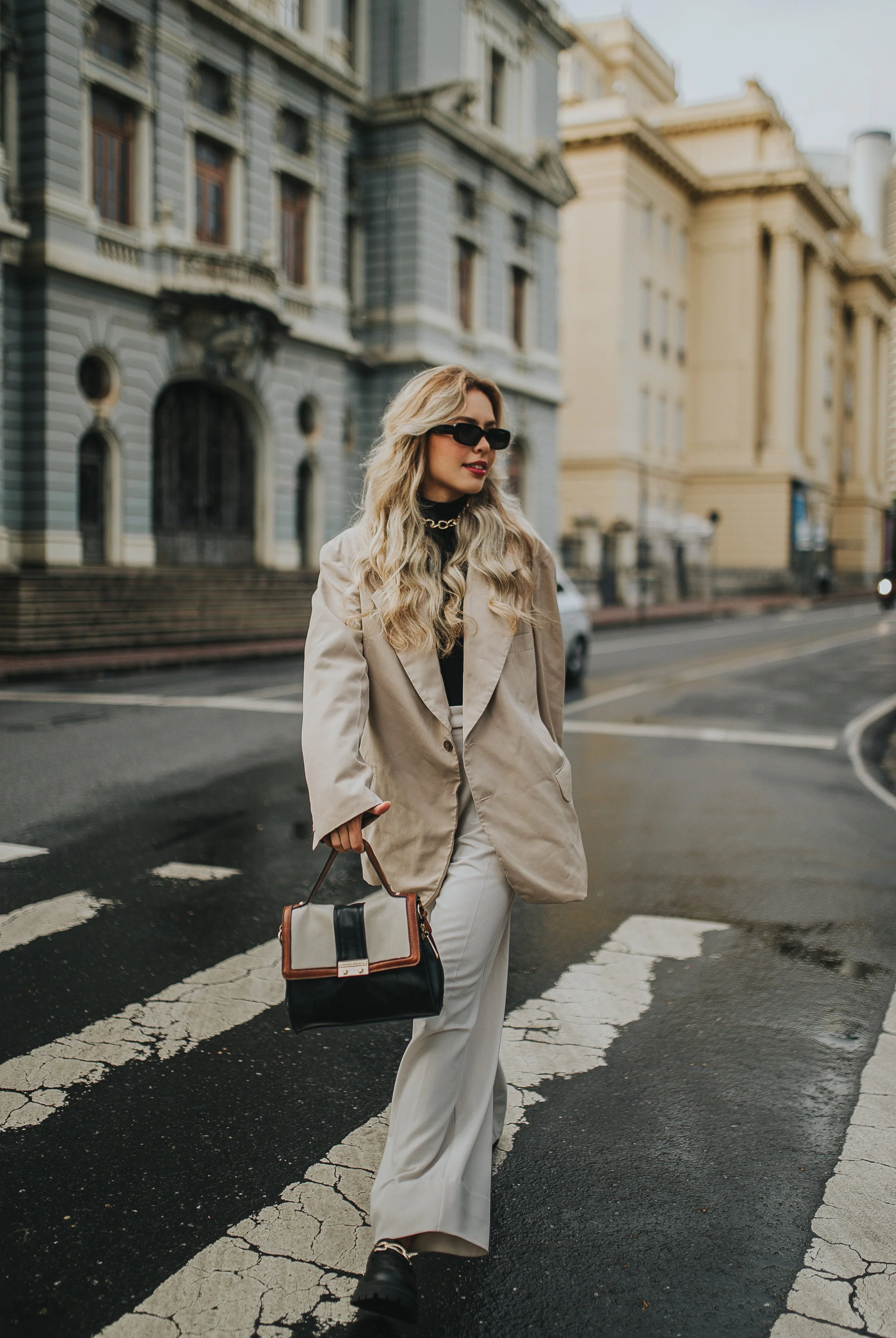 A woman with long blonde hair wearing a beige trench coat, black sunglasses, and black boots crossing a street with city buildings in the background during daytime.