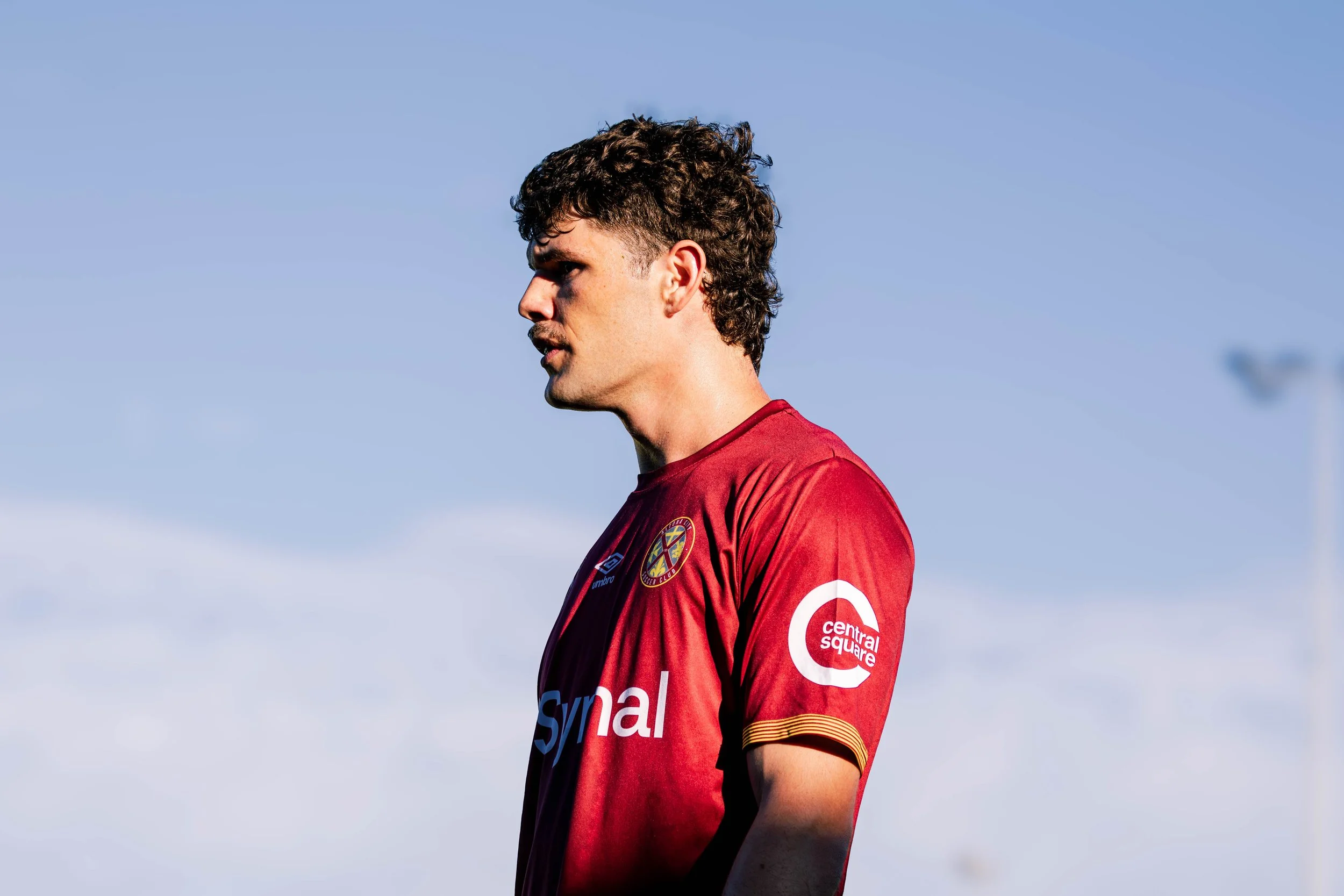 Side profile of a young man with curly hair wearing a red sports jersey with logos and standing against a blue sky.