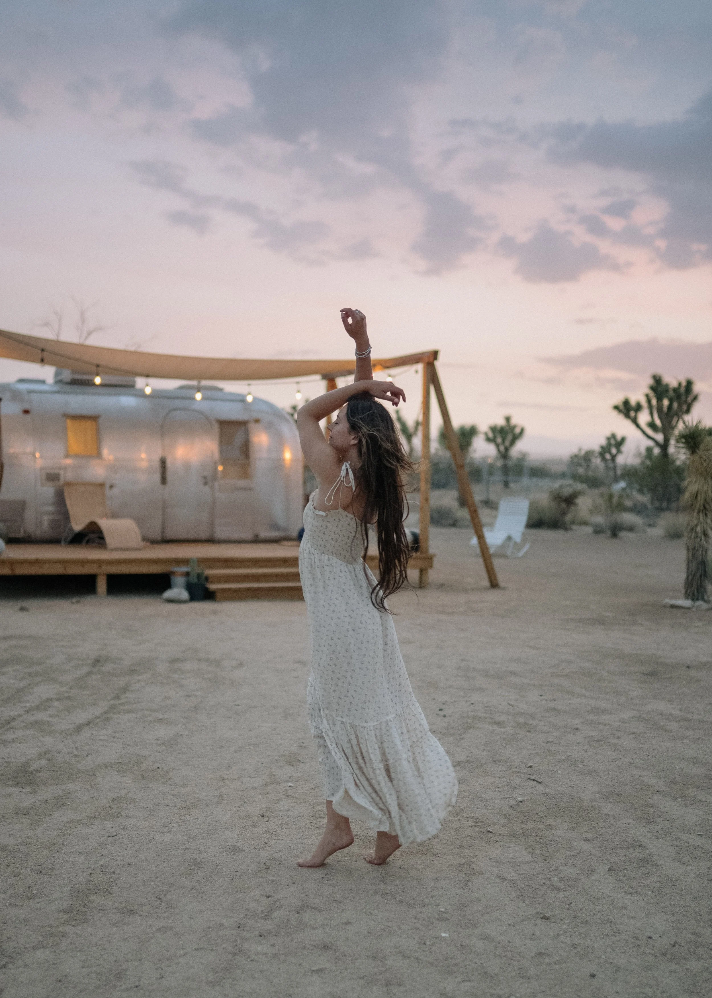 A woman in a white dress dancing barefoot on a sandy desert at sunset, with a vintage camper trailer and wooden swing set in the background.