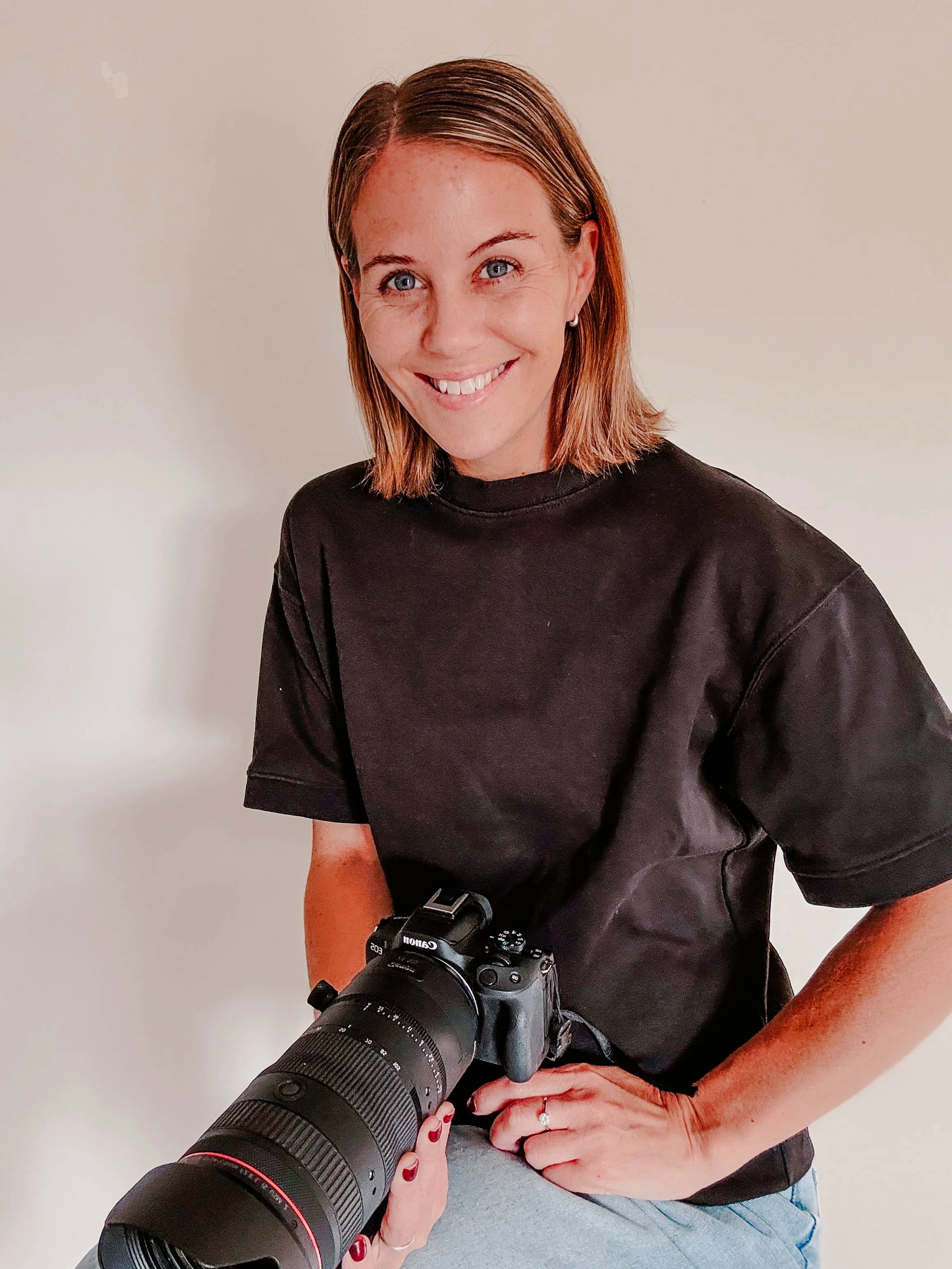 A woman with shoulder-length brown hair, blue eyes, and fair skin smiling while holding a professional camera with a large telephoto lens. She is wearing a black t-shirt and has red nail polish, with a light gray background.