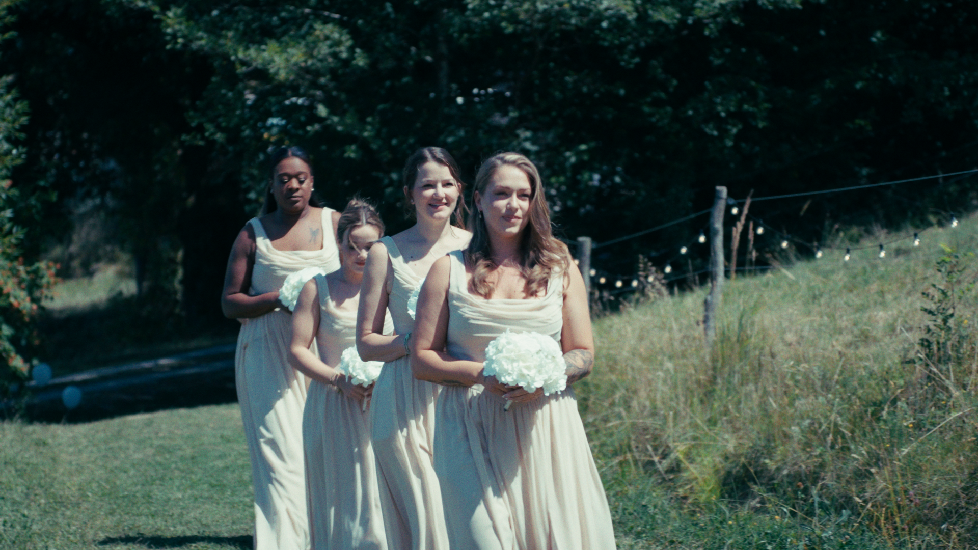 Group of bridesmaids in cream-colored dresses holding white flower bouquets, walking outdoors along a grass path with trees and a fence in the background.