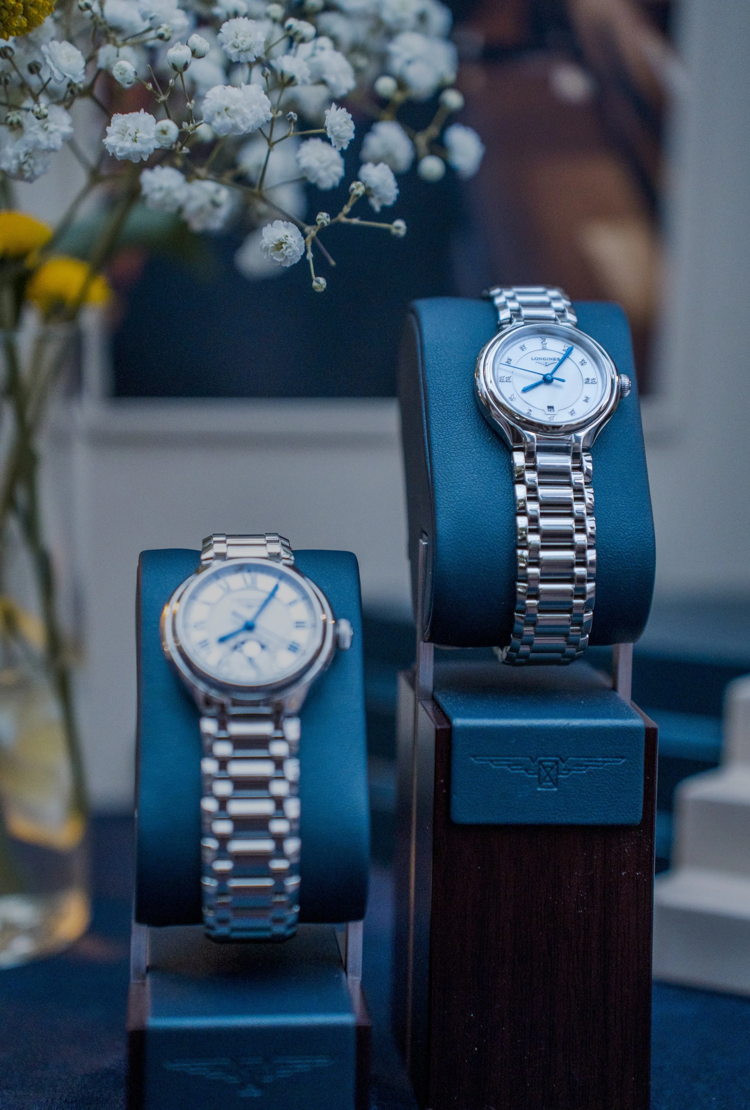 Three luxury watches on display, each on a stand, with white flowers in the foreground.