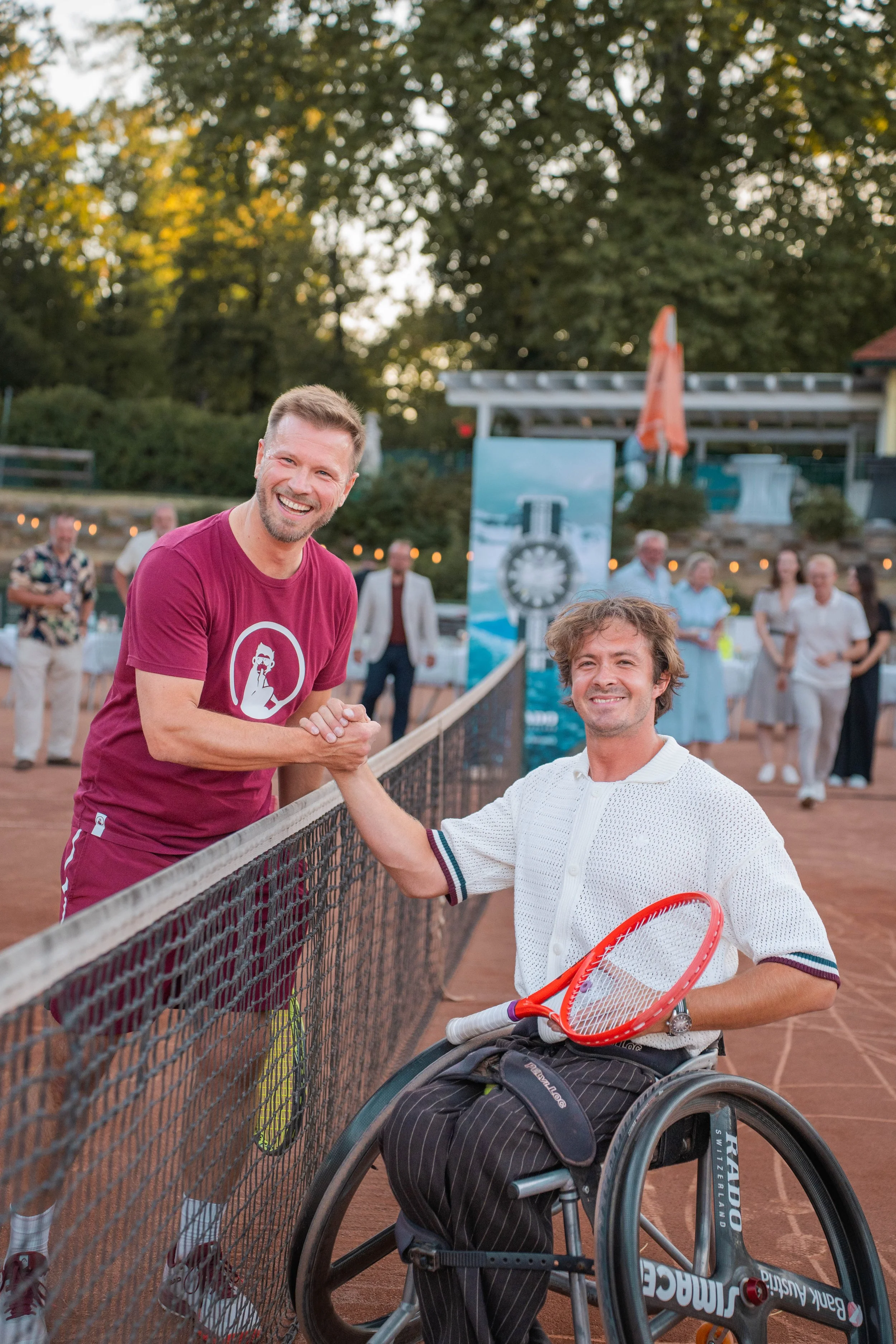 Two men shaking hands on a tennis court, with one in a wheelchair holding a tennis racket, smiling, during sunset with people in the background.